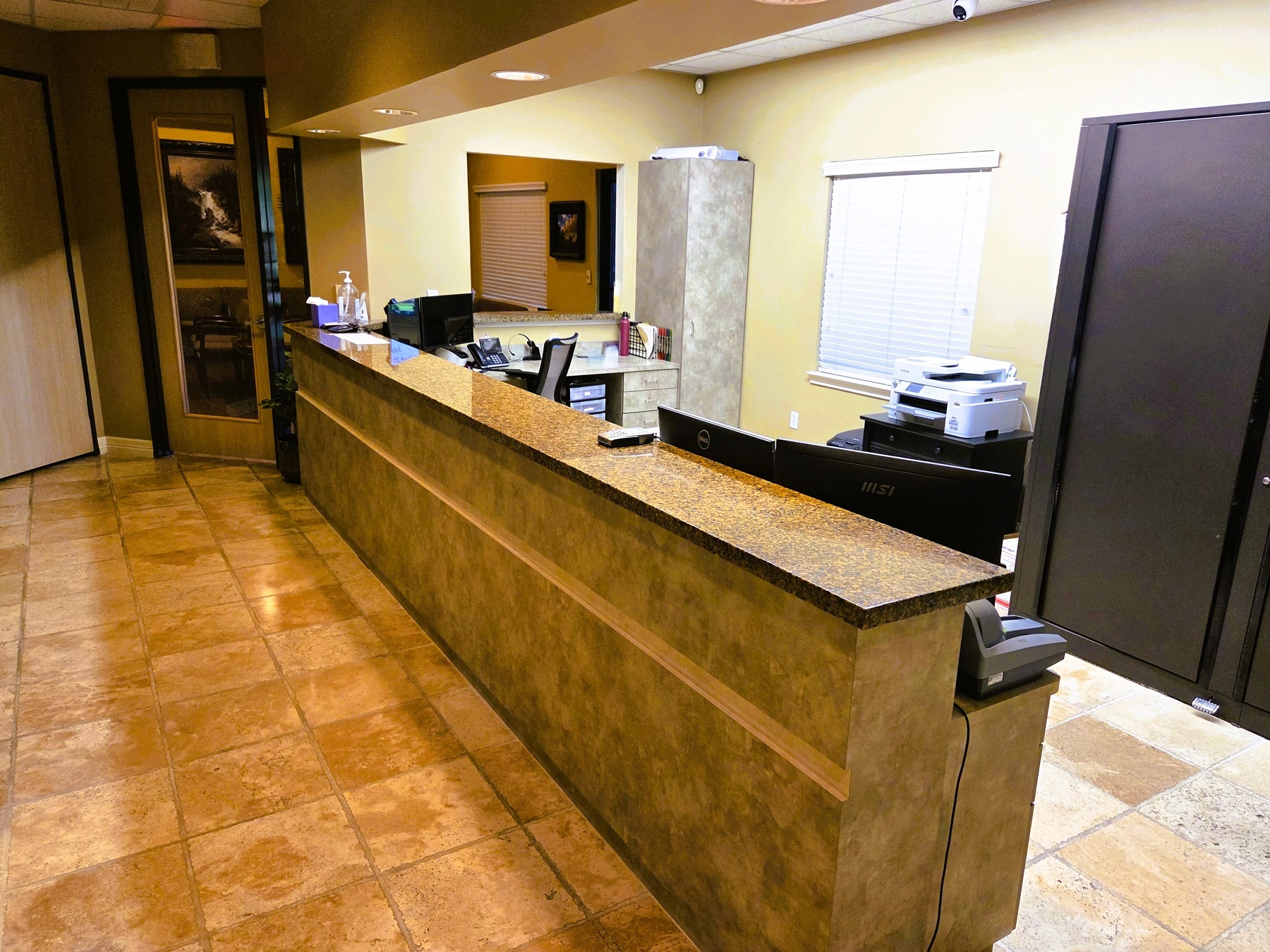 Light tan walls with a long builtin countertop and large terracotta tile flooring. There's a front office area behind the countertop. There's rolling office chairs and equipment.