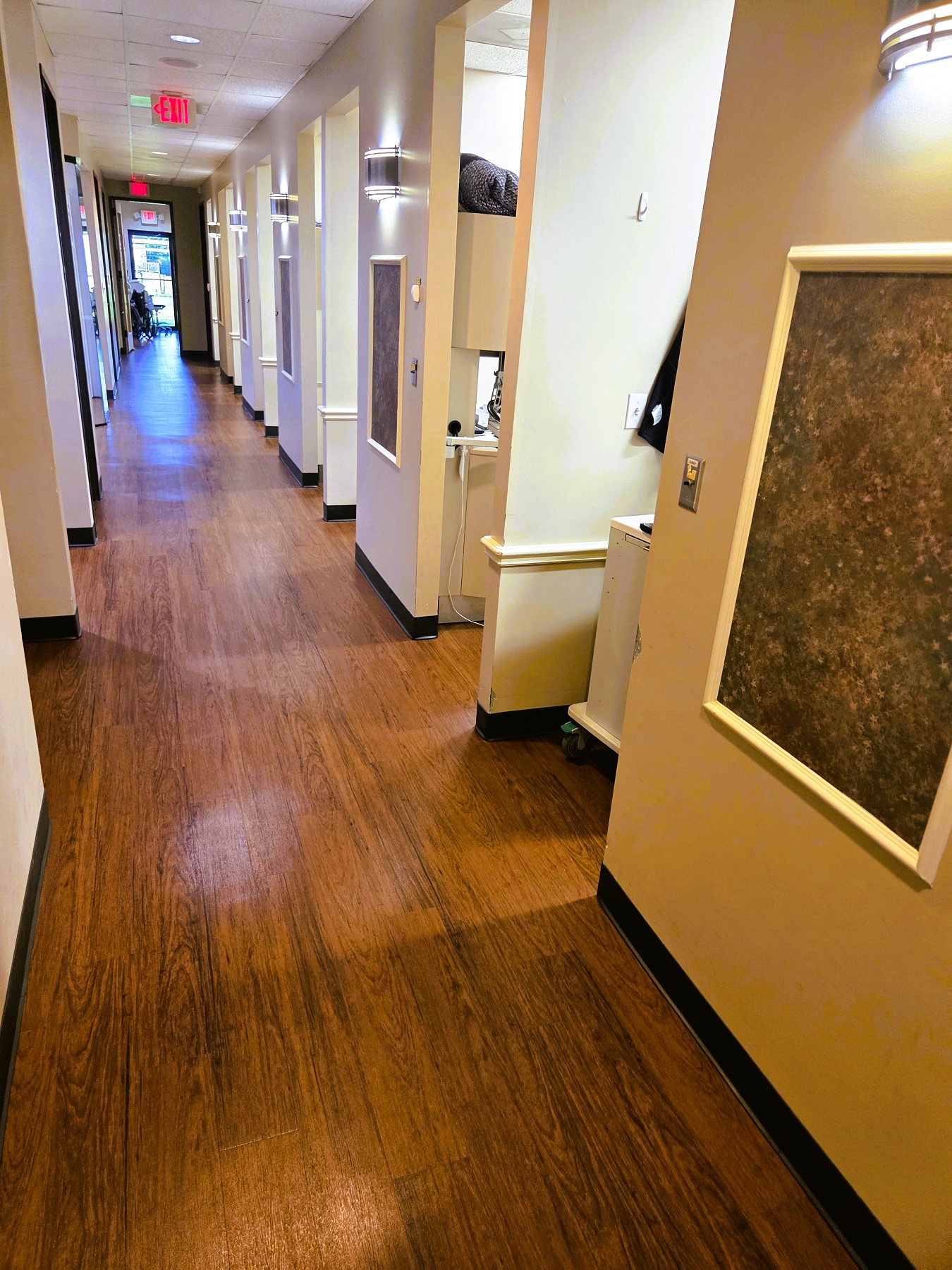Beige walls line a long hallway with entry ways to individual operatories and rooms. There are dark brown squares with white frames and lighted wall sconces between each entry way. Wood-look vinyl flooring runs the length of the hallway.