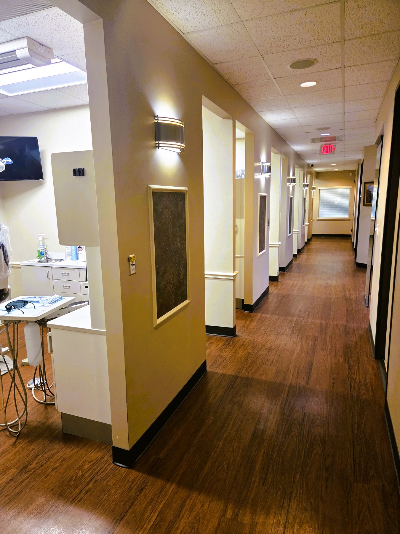 Beige walls line a long hallway with entry ways to individual operatories and rooms. There are dark brown squares with white frames and lighted wall sconces between each entry way. Wood-look vinyl flooring runs the length of the hallway.