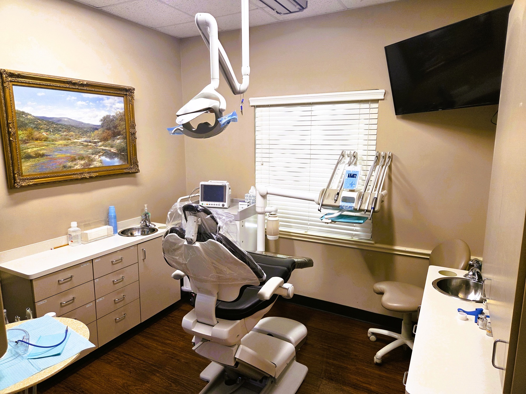 Beige walls and cabinetry with white countertops and stainless steel sink. Large framed artwork on the wall. A dental procedural chair anchors the room with overhead dental lighting and rolling dental stools and equipment trays. There's lots of space for tools,, equipment and supplies.
