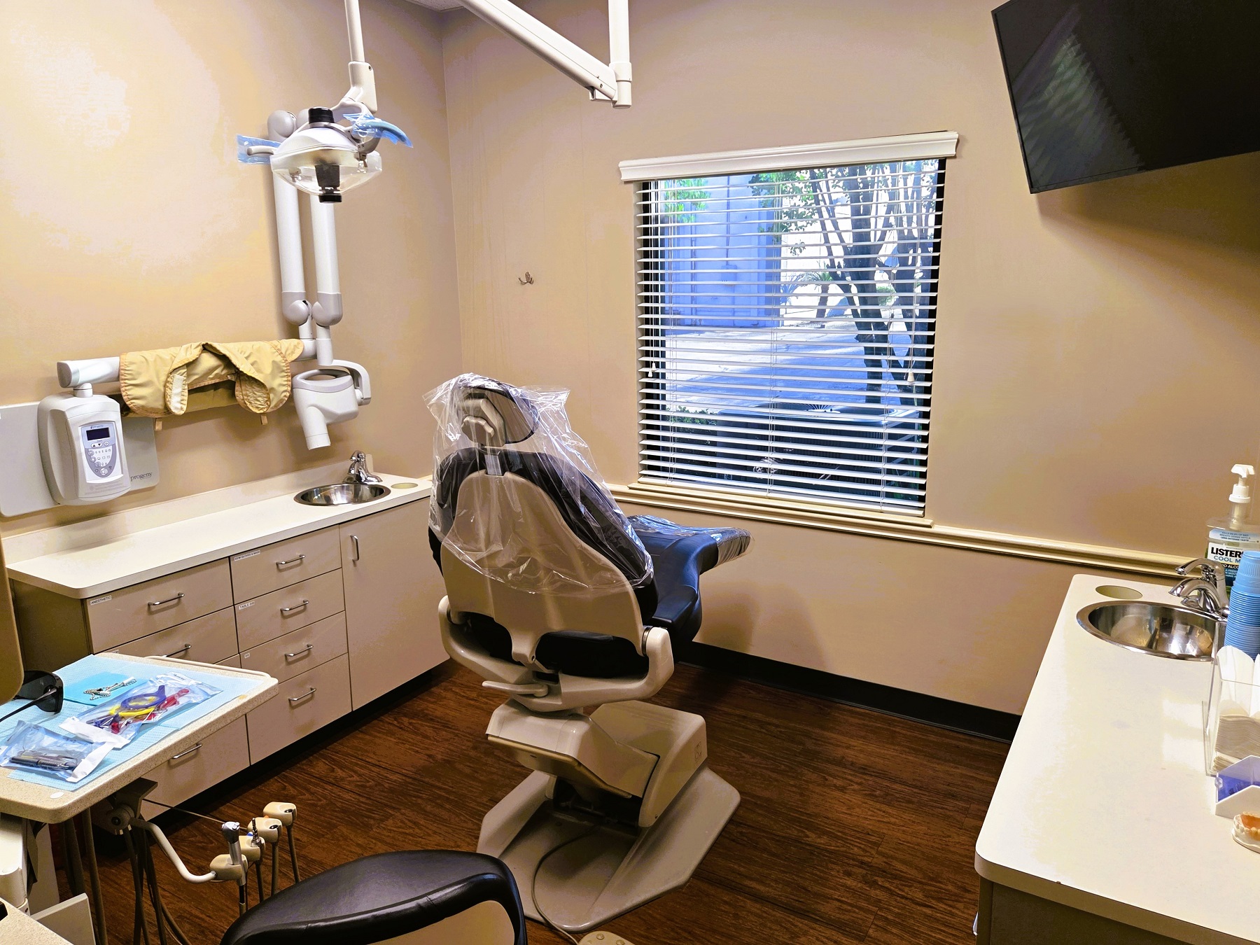 Beige walls and cabinetry with white countertops and stainless steel sink. Large framed artwork on the wall. A dental procedural chair anchors the room with overhead dental lighting and rolling dental stools and equipment trays. There's lots of space for tools,, equipment and supplies.