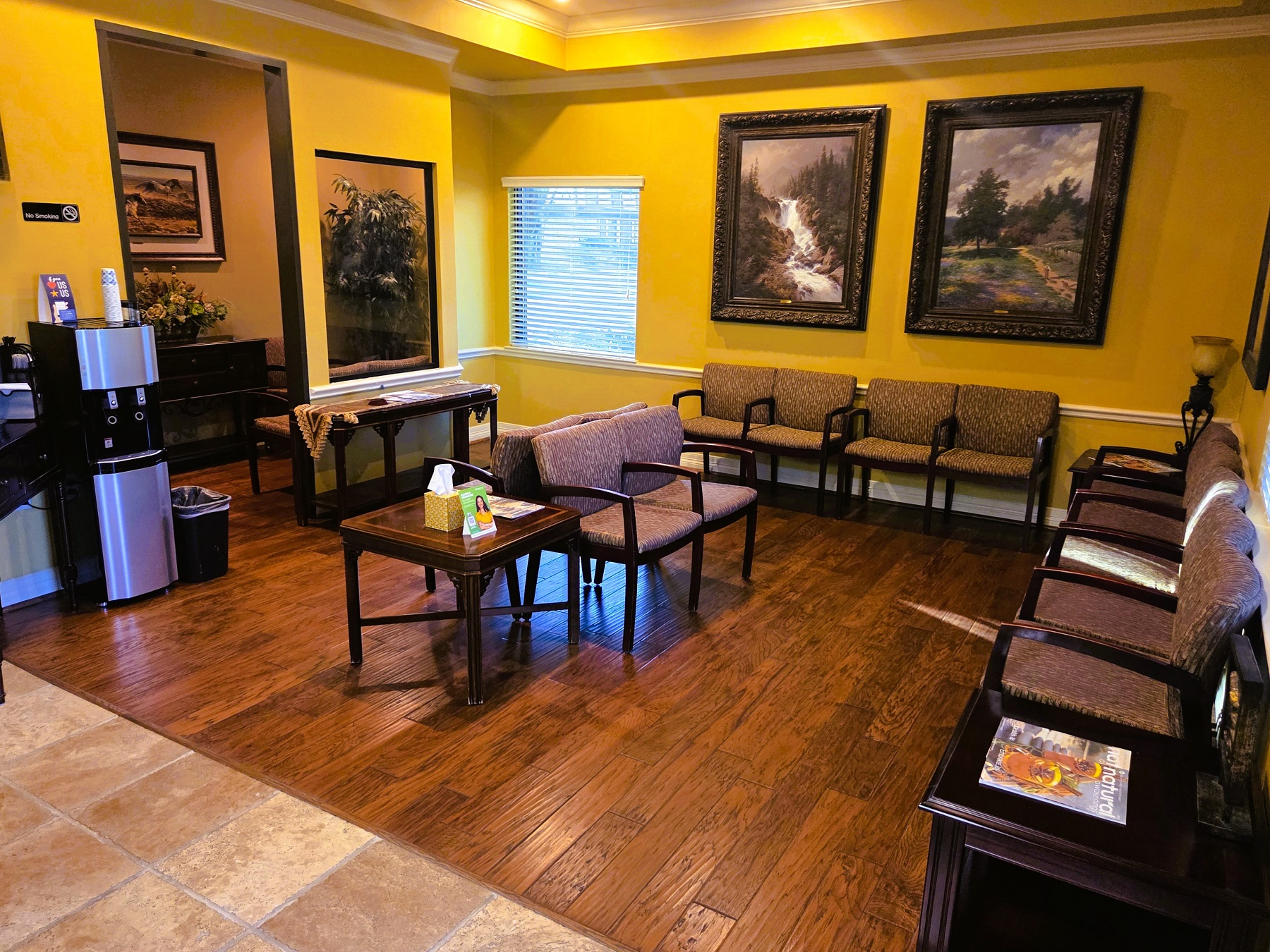 Light yellow walls with a white chair rail, baseboard, and crown molding. A large window lets in lots of natural light. Wood-look vinyl flooring provides warmth. The waiting area in furnished with cushioned, wood arm chairs. There's a drink station and the flooring transitions to large terracotta tiles. Lots of framed artwork ads warmth to the waiting area.