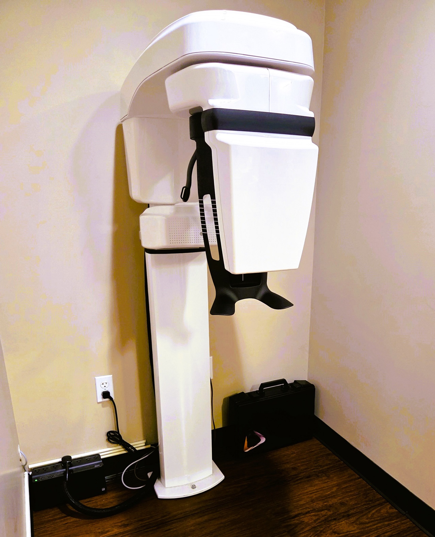 Beige walls surround a large dental x-ray machine standing against the wall with commercial carpet tile flooring.