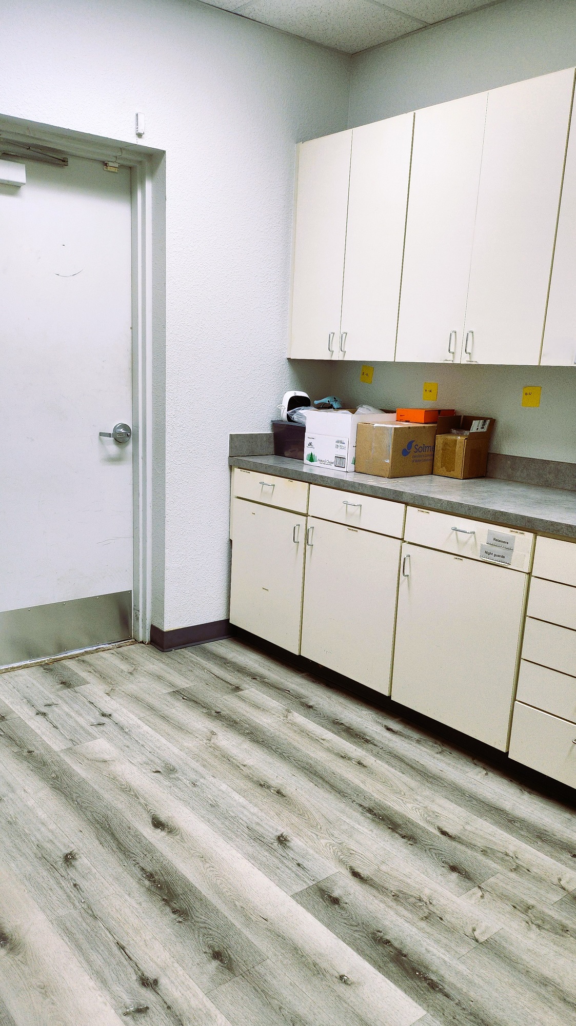 Grey walls with dark baseboards and grey wood-look vinyl flooring. White uppter and lower cabinets hold lots of dental lab equipment and supplies.