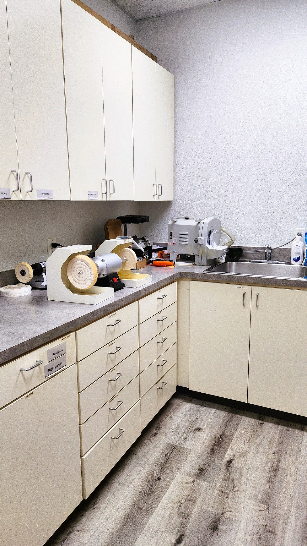 Grey walls with dark baseboards and grey wood-look vinyl flooring. White uppter and lower cabinets hold lots of dental lab equipment and supplies.
