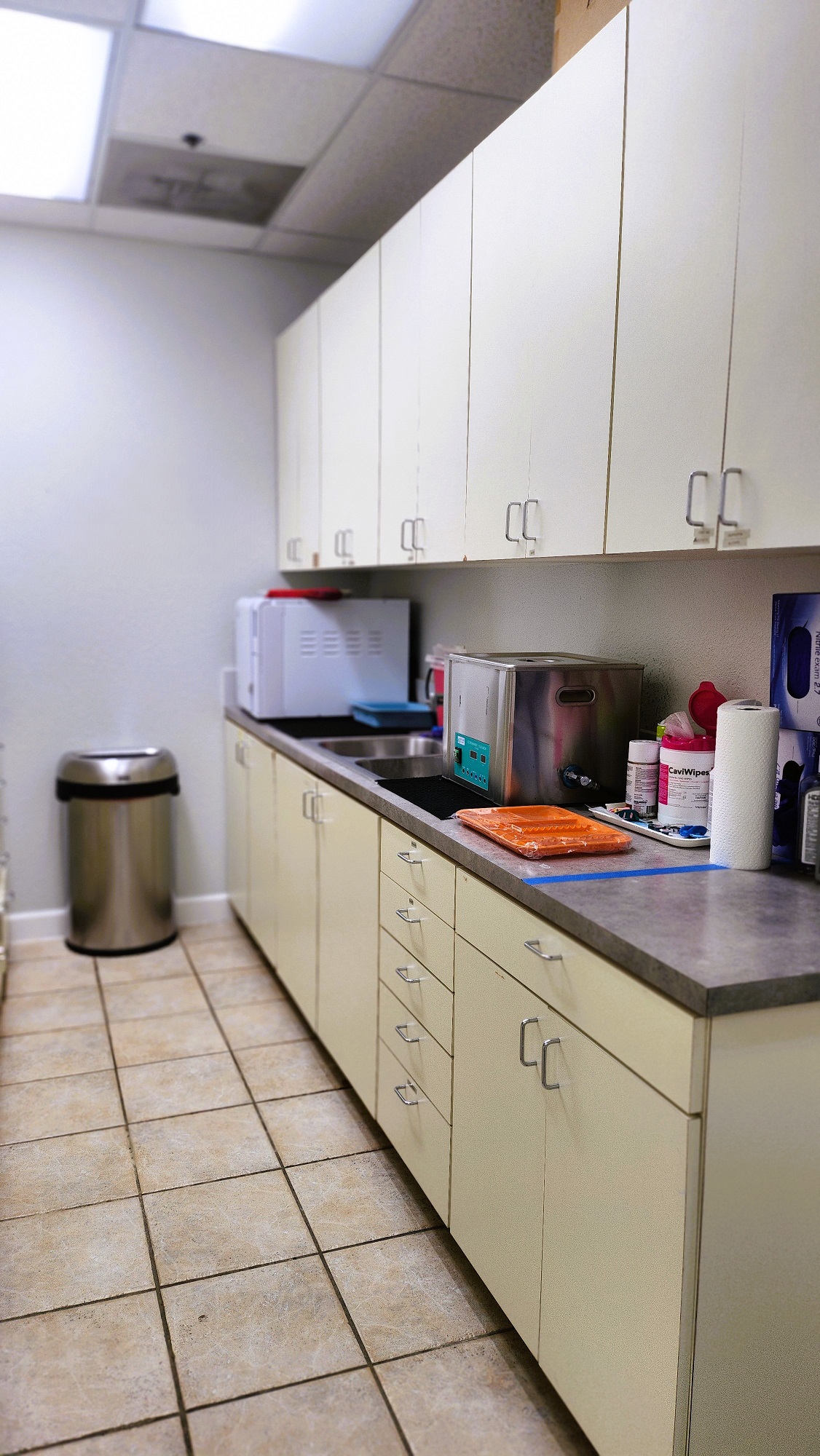 Light grey walls with large tan tile flooring. White upper and lower cabinets with grey counter tops hold lots of dental sterilization supplies. A stainless steel trash can sits in the back along the wall.