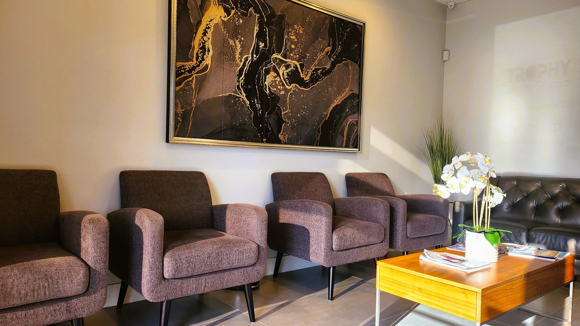 Light grey walls with dark baseboards and medium colored carpet. The walls are lined with overstuffed arm chairs. A large piece of framed artwork sits on the wall. There's a coffee table in the center of the room with a matching end table in the corner holding up a snake plant.