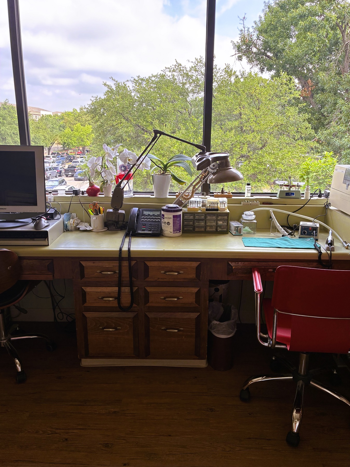 Huge window looks out on the treetops. A builtin desk and counter top holds tons of dental lab equipment, supplies, and tools. An orange office chair rolls around on hardwood floors.