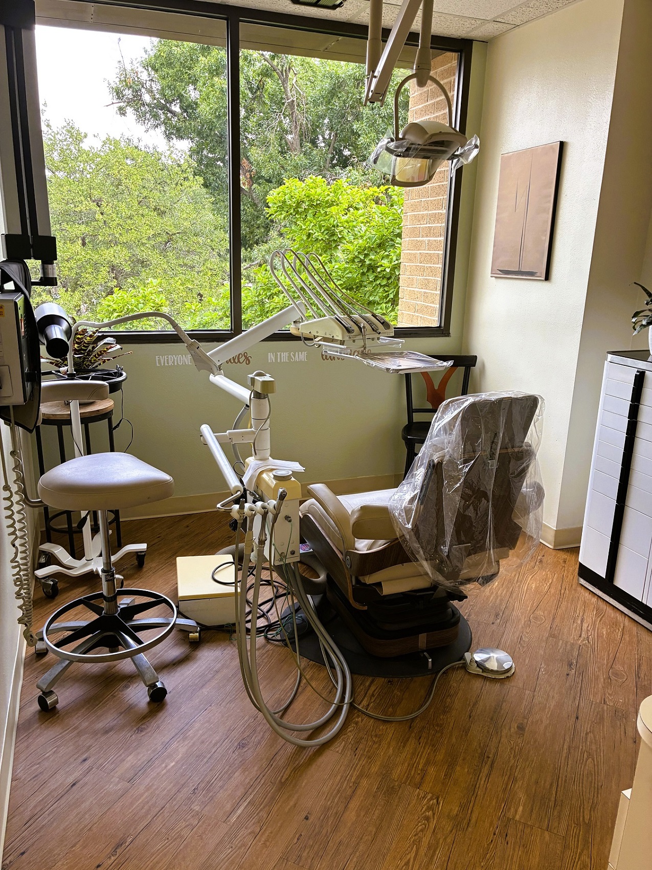 Light walls with a huge picture window looking out onto the tree tops. Hardwood flooring with a light dental procedural chair anchoring the room with overhead dental lighting. Rolling dental stool and carts surround the chair with lots of dental tools and equipment. The room is flooded with lots of natural light.