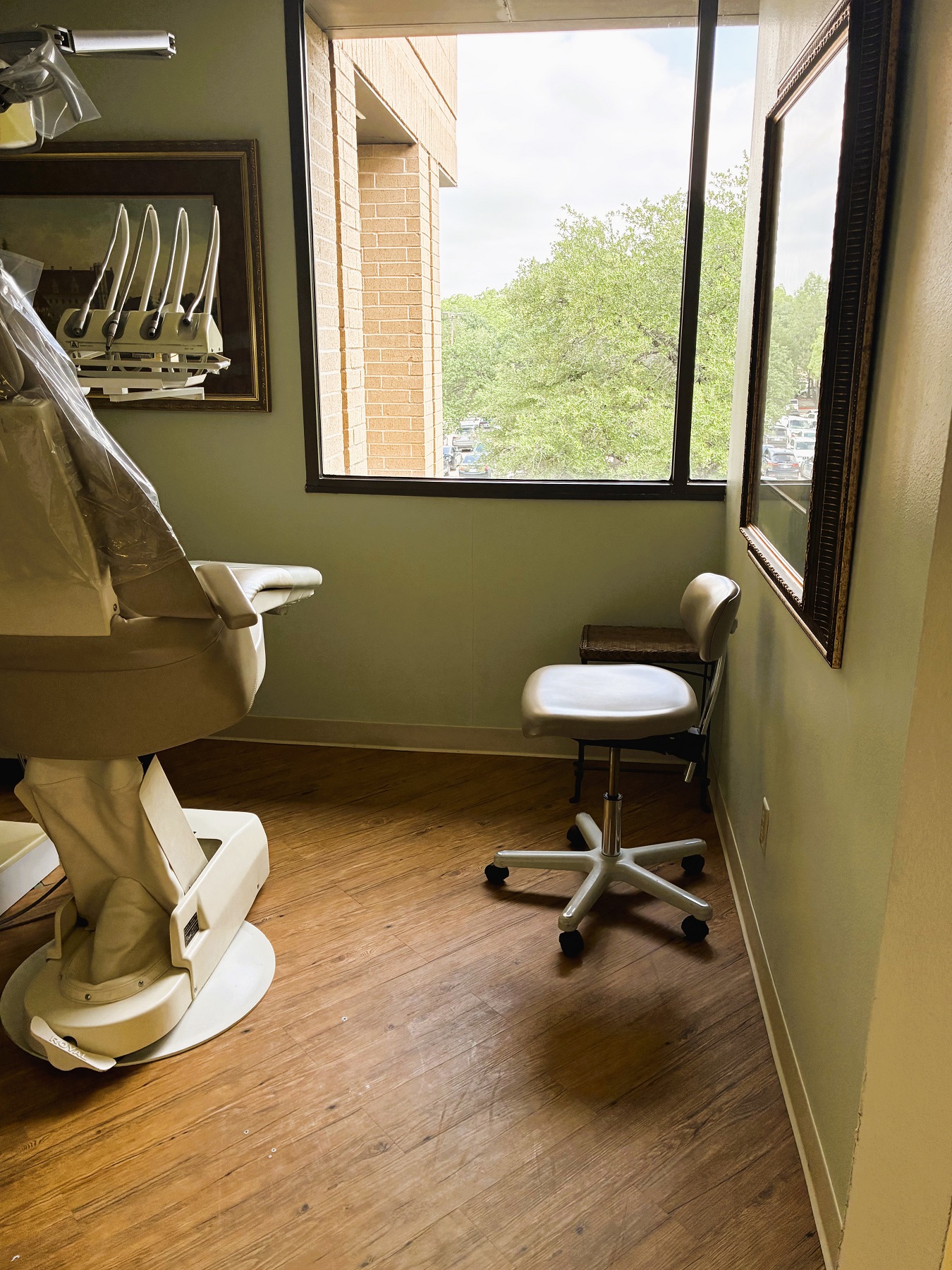 Light walls with a huge picture window looking out onto the tree tops. Hardwood flooring with a light dental procedural chair anchoring the room with overhead dental lighting. Rolling dental stool and carts surround the chair with lots of dental tools and equipment. The room is flooded with lots of natural light.