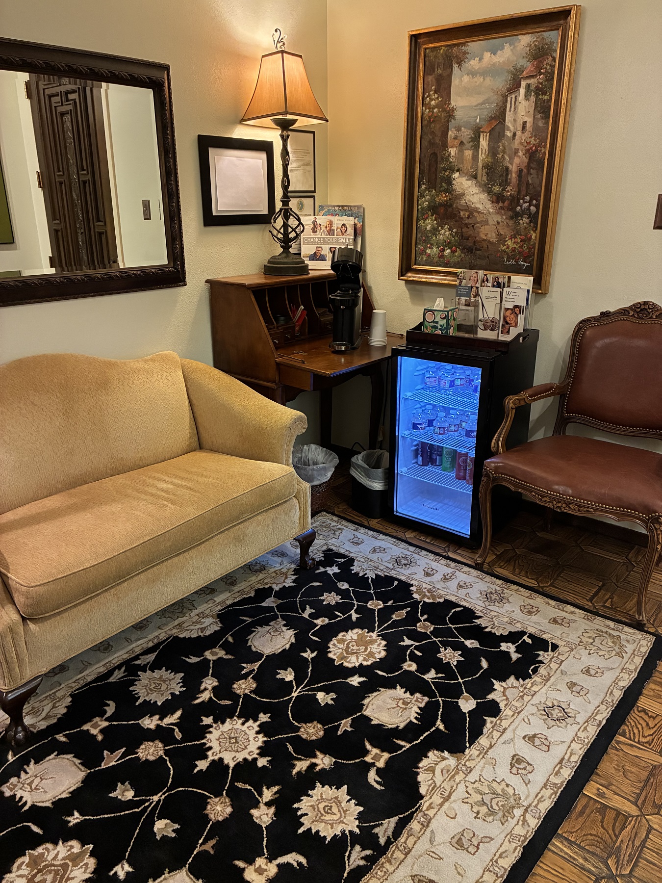 Light walls with framed art and mirrors hanging on them. Vintage sofa's line the wall and surround a rug that covers hardwood flooring. There is a mini fridge with cold beverages for waiting patients and a side table holding a reading lamp and reading material.