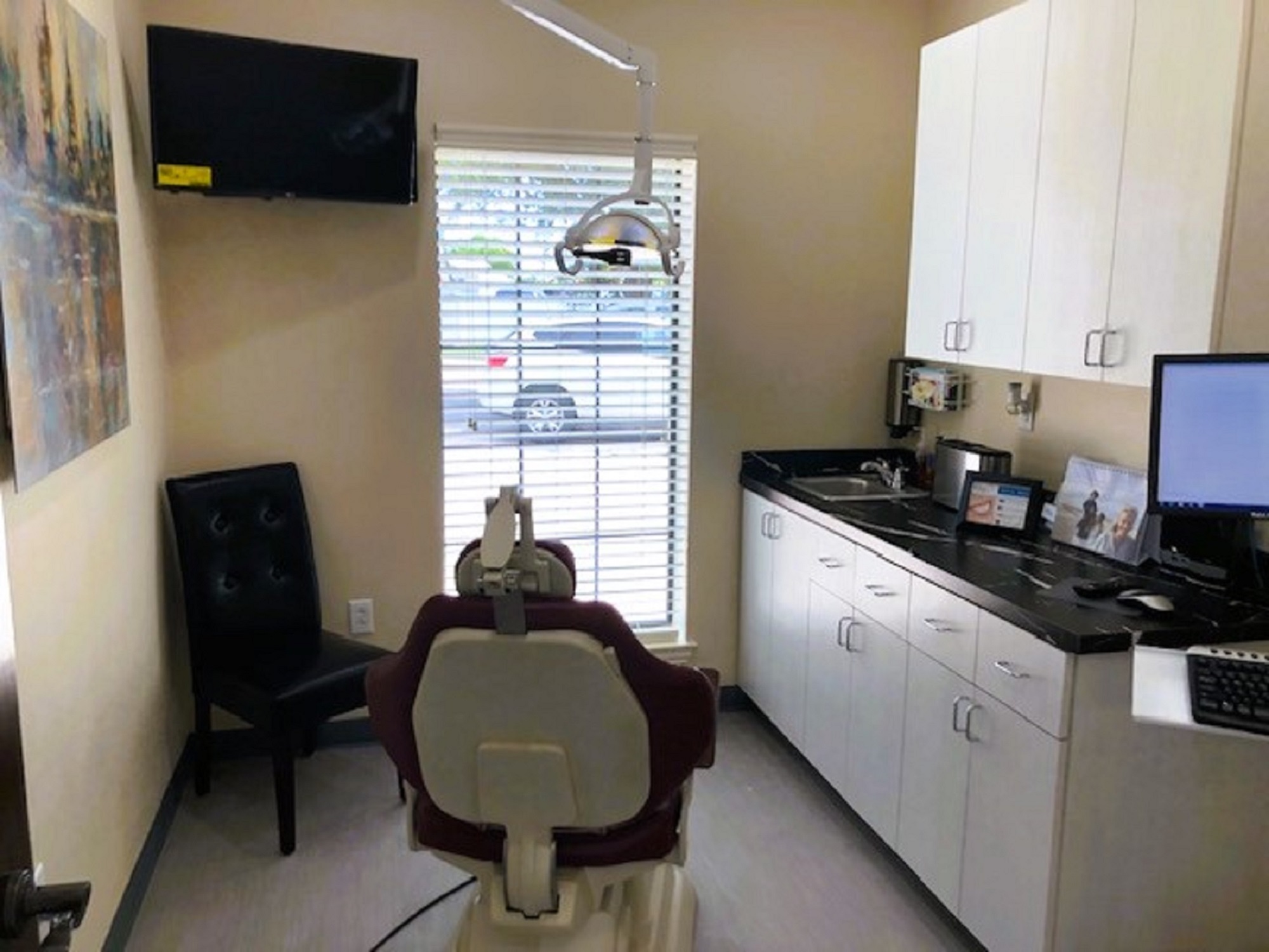 Beige walls with black base boards. A dark cushioned chair sits in the corner. A tall window sits between it and the cabinet that runs the full length of the wall, letting in lots of natural light. A dental examination chair anchors the room. An off white cabinet, both upper and lower, runs full length of the wall with a dark countertop. A large piece of artwork hangs on the opposite wall.