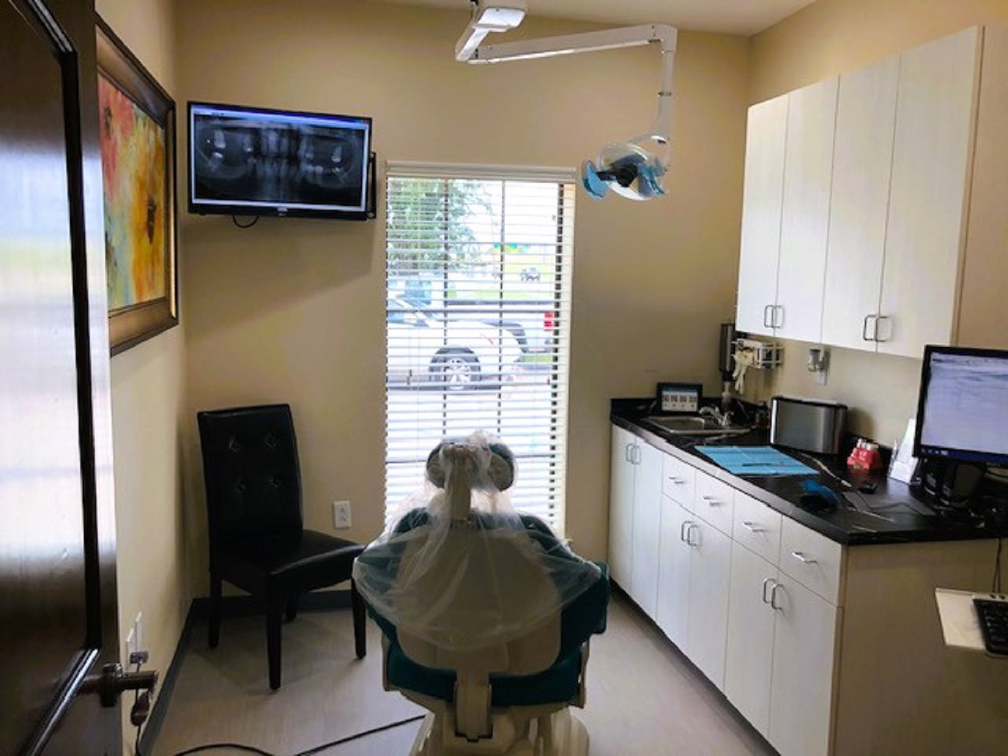 Beige walls with black base boards. A dark cushioned chair sits in the corner. A tall window sits between it and the cabinet that runs the full length of the wall, letting in lots of natural light. A dental examination chair anchors the room. An off white cabinet, both upper and lower, runs full length of the wall with a dark countertop. A large piece of artwork hangs on the opposite wall.