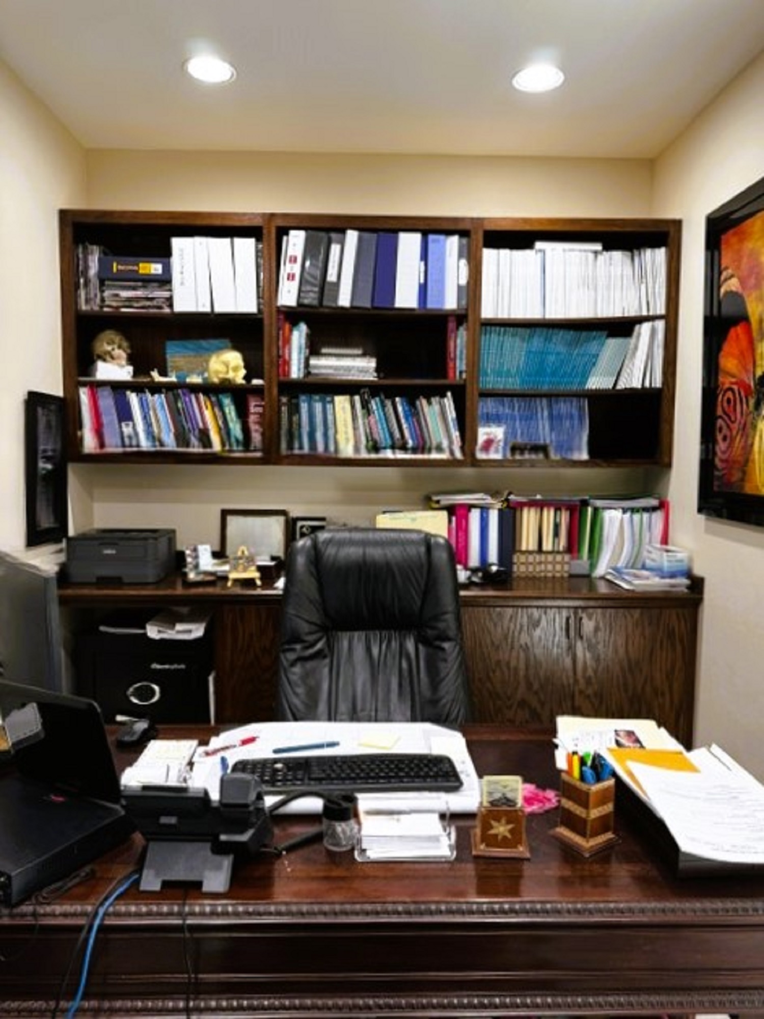 Beige walls. Mahogany stained executive desk with matching wall desk and book shelves behind it. A large black executive rolling chair sits between them. The shelves are filled with book and the desk has a computer with keyboard and monitor and some file sitting on it.