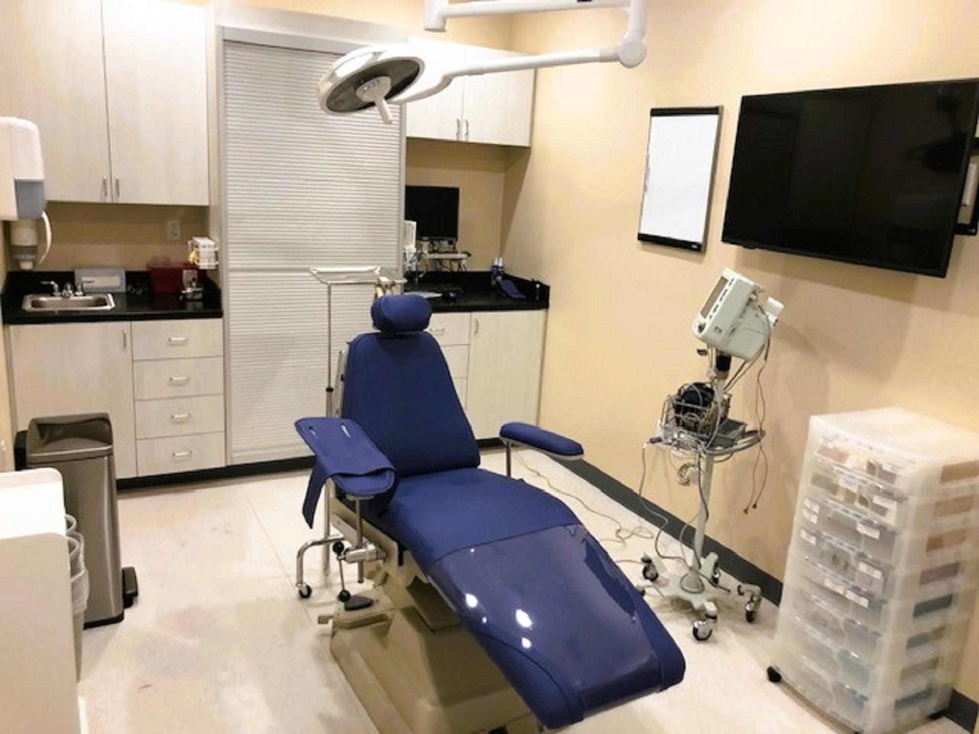 Beige walls with black base boards and beige tile flooring. A dental exam chair anchors the room. Off white cabinetry covers one entire wall with black counter tops and holds lots of dental tools, equipment and supplies. An overhead dental light is over the exam chair. Several rolling carts hold additional supplies.