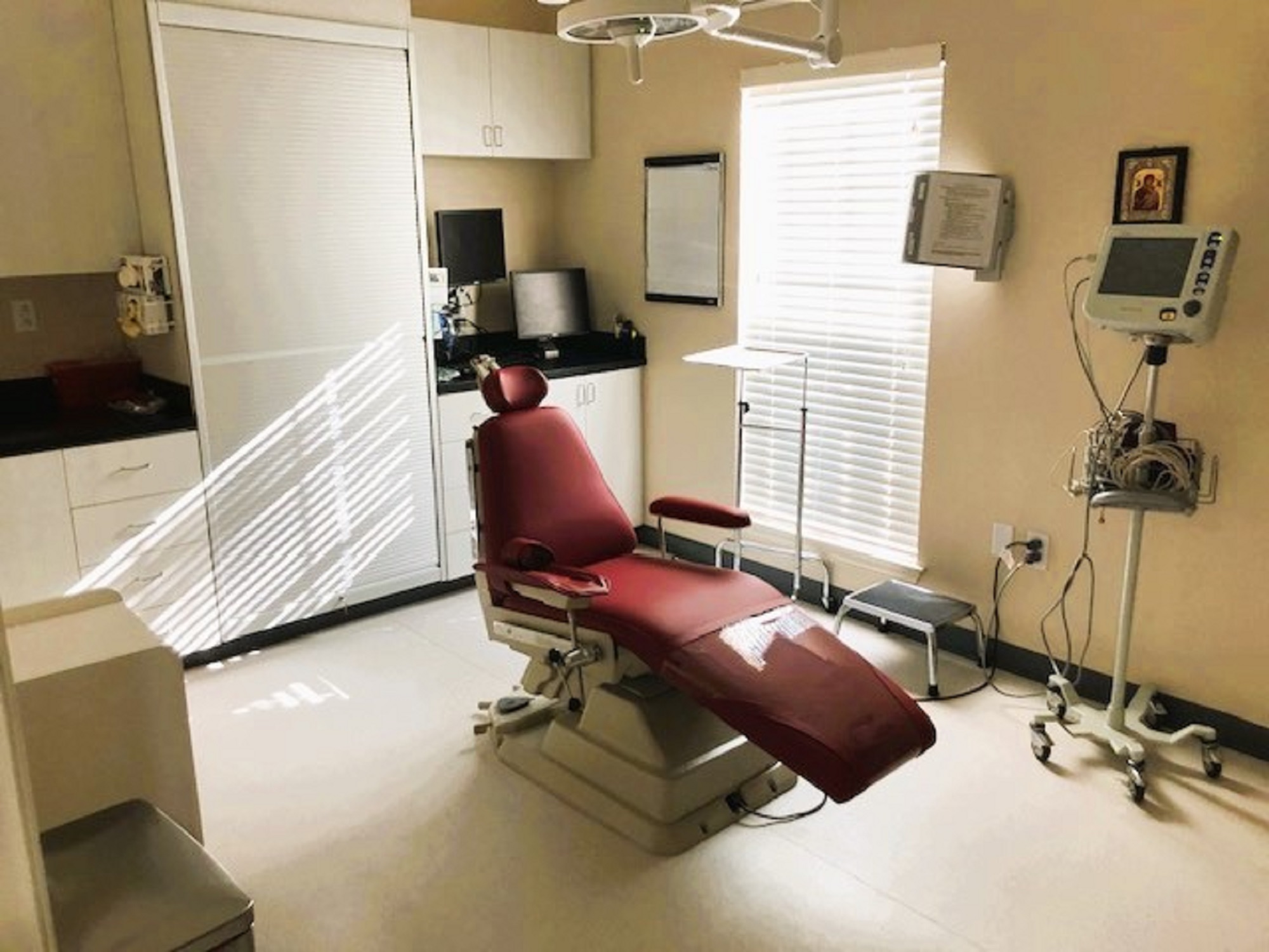Beige walls with black base boards and beige tile flooring. A dental exam chair anchors the room. Off white cabinetry covers one entire wall with black counter tops and holds lots of dental tools, equipment and supplies. An overhead dental light is over the exam chair. Several rolling carts hold additional supplies.