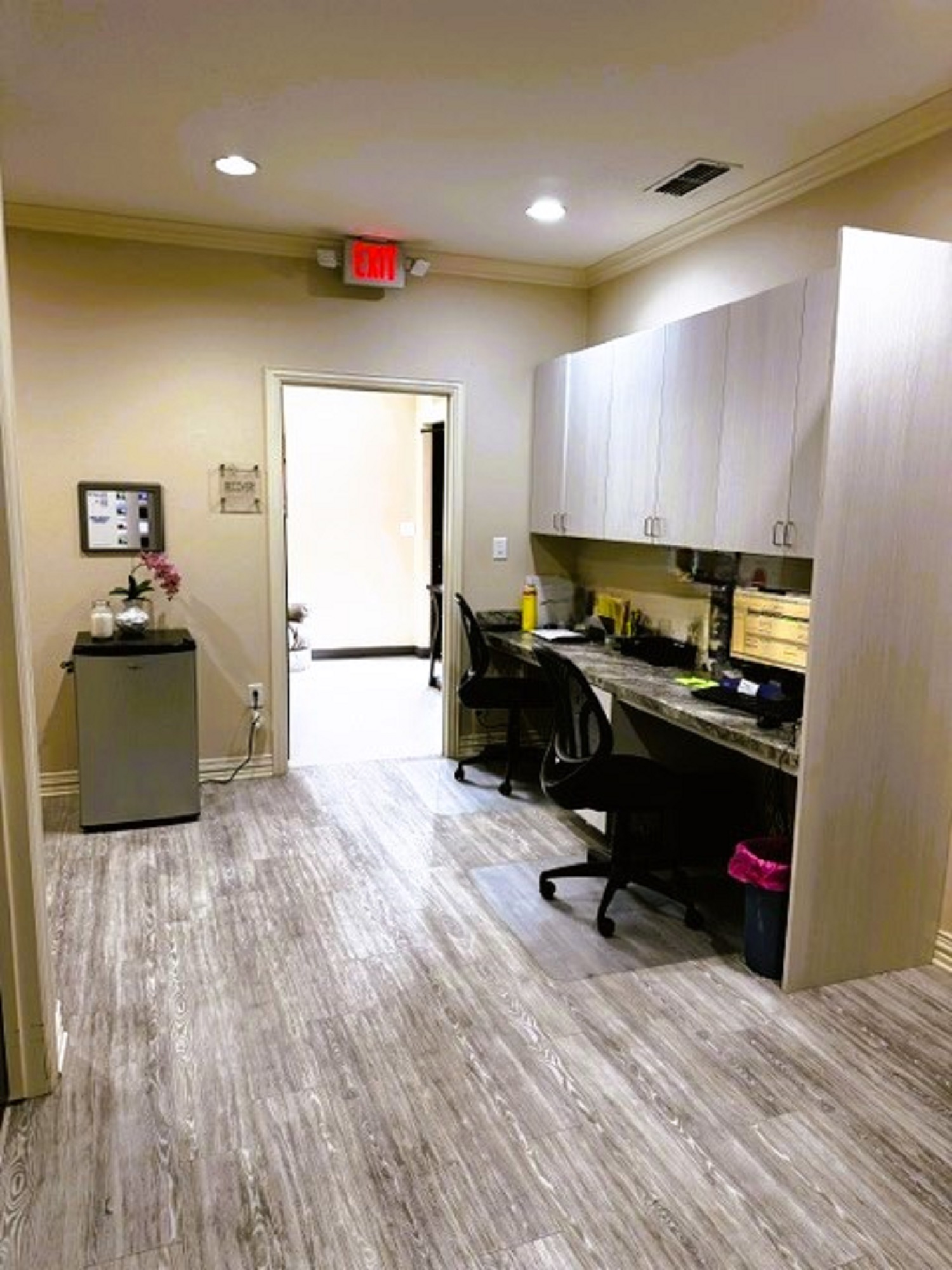 Beige walls with cream trim and grey wood-look flooring. A two desk built in runs the length on one wall with Upper and lower cabinetys and a full length desk top. Two rolling office chairs. Lights under the upper cabinets to light the desk top.