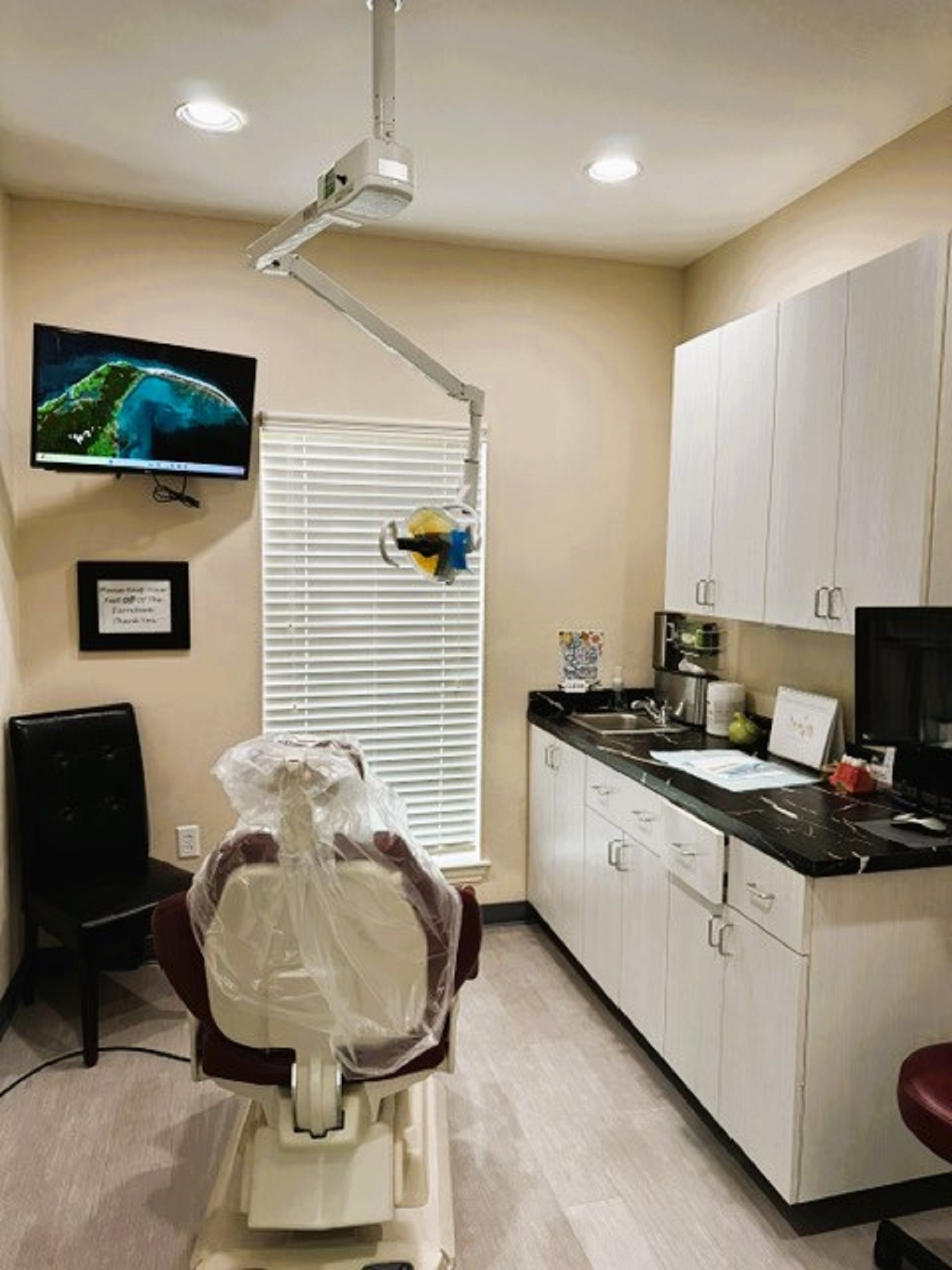 Beige walls with black base boards and beige tile flooring. A dental exam chair anchors the room. Off white cabinetry covers one entire wall with black counter tops and holds lots of dental tools, equipment and supplies. An overhead dental light is over the exam chair. Several rolling carts hold additional supplies.