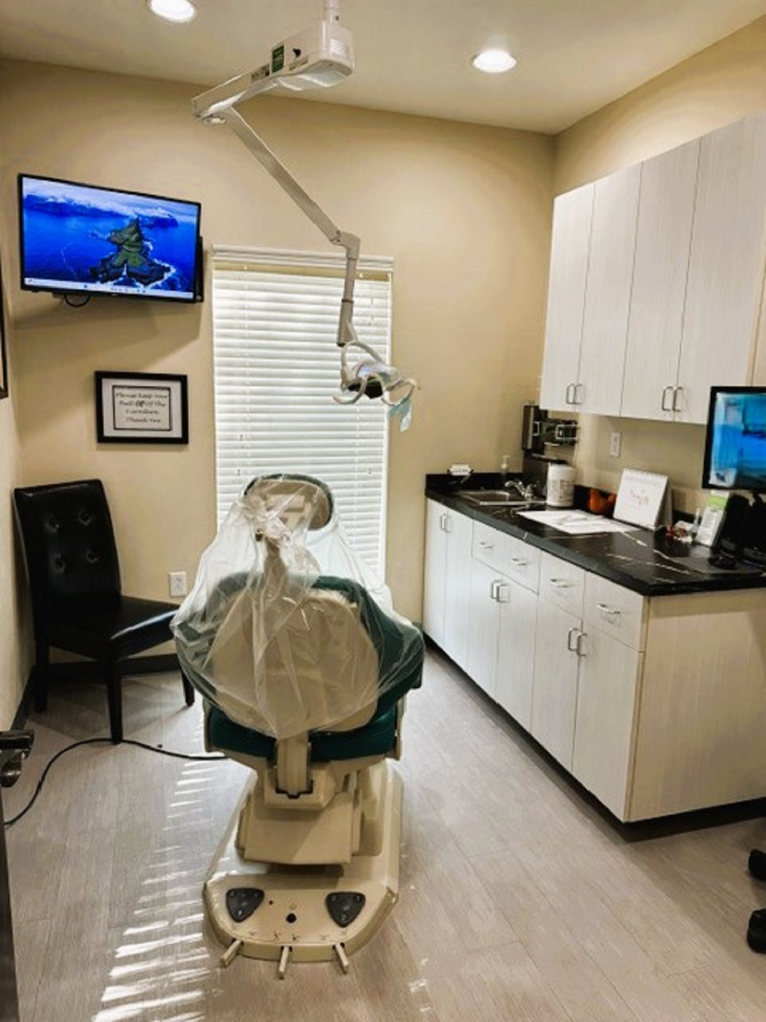 Beige walls with black base boards and beige tile flooring. A dental exam chair anchors the room. Off white cabinetry covers one entire wall with black counter tops and holds lots of dental tools, equipment and supplies. An overhead dental light is over the exam chair. Several rolling carts hold additional supplies.