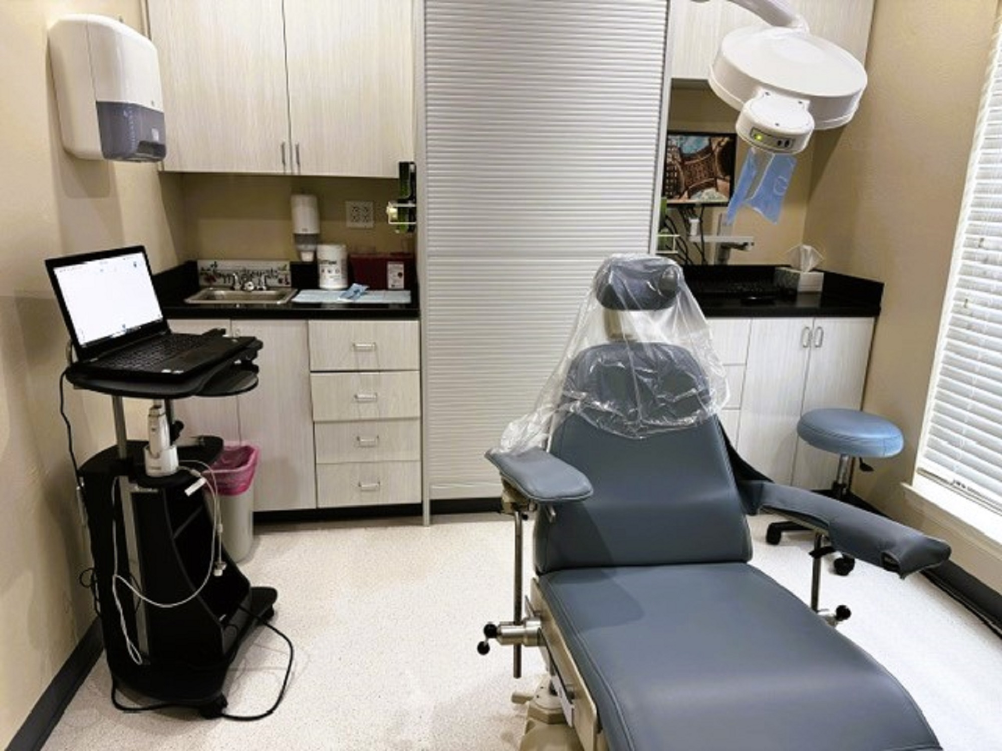 Beige walls with black base boards and beige tile flooring. A dental exam chair anchors the room. Off white cabinetry covers one entire wall with black counter tops and holds lots of dental tools, equipment and supplies. An overhead dental light is over the exam chair. Several rolling carts hold additional supplies.