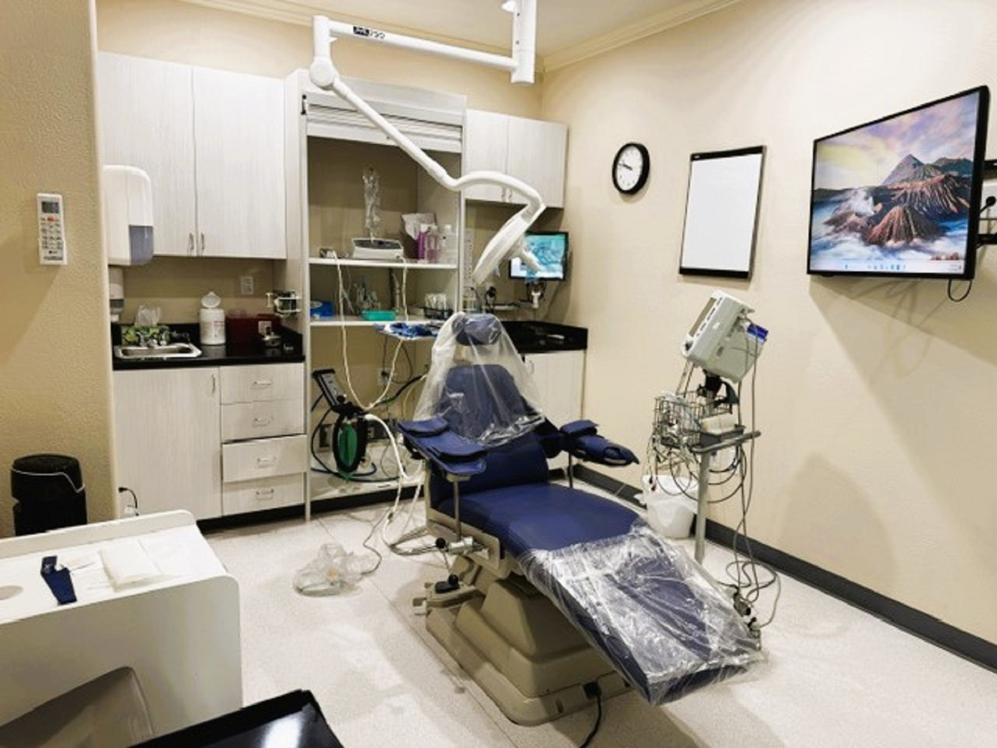 Beige walls with black base boards and beige tile flooring. A dental exam chair anchors the room. Off white cabinetry covers one entire wall with black counter tops and holds lots of dental tools, equipment and supplies. An overhead dental light is over the exam chair. Several rolling carts hold additional supplies.