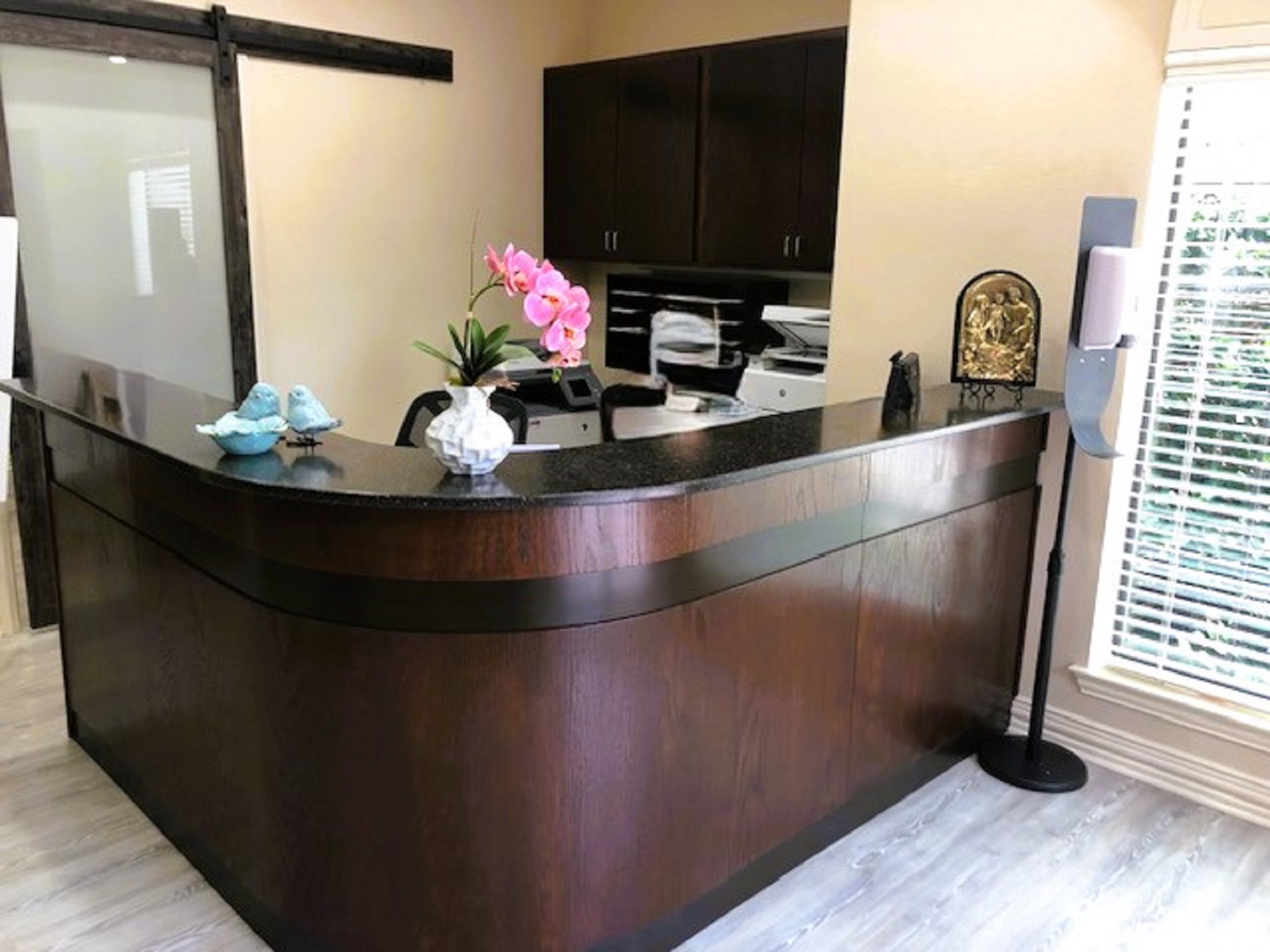 Beige walls with cream base boards. A black framed barn door sits behind the large "L" shaped, darkly stained wood desk with a black stone countertop. Matching dark cabinetry is hung on the wall behind the desk. A black office chair sits behind the desk