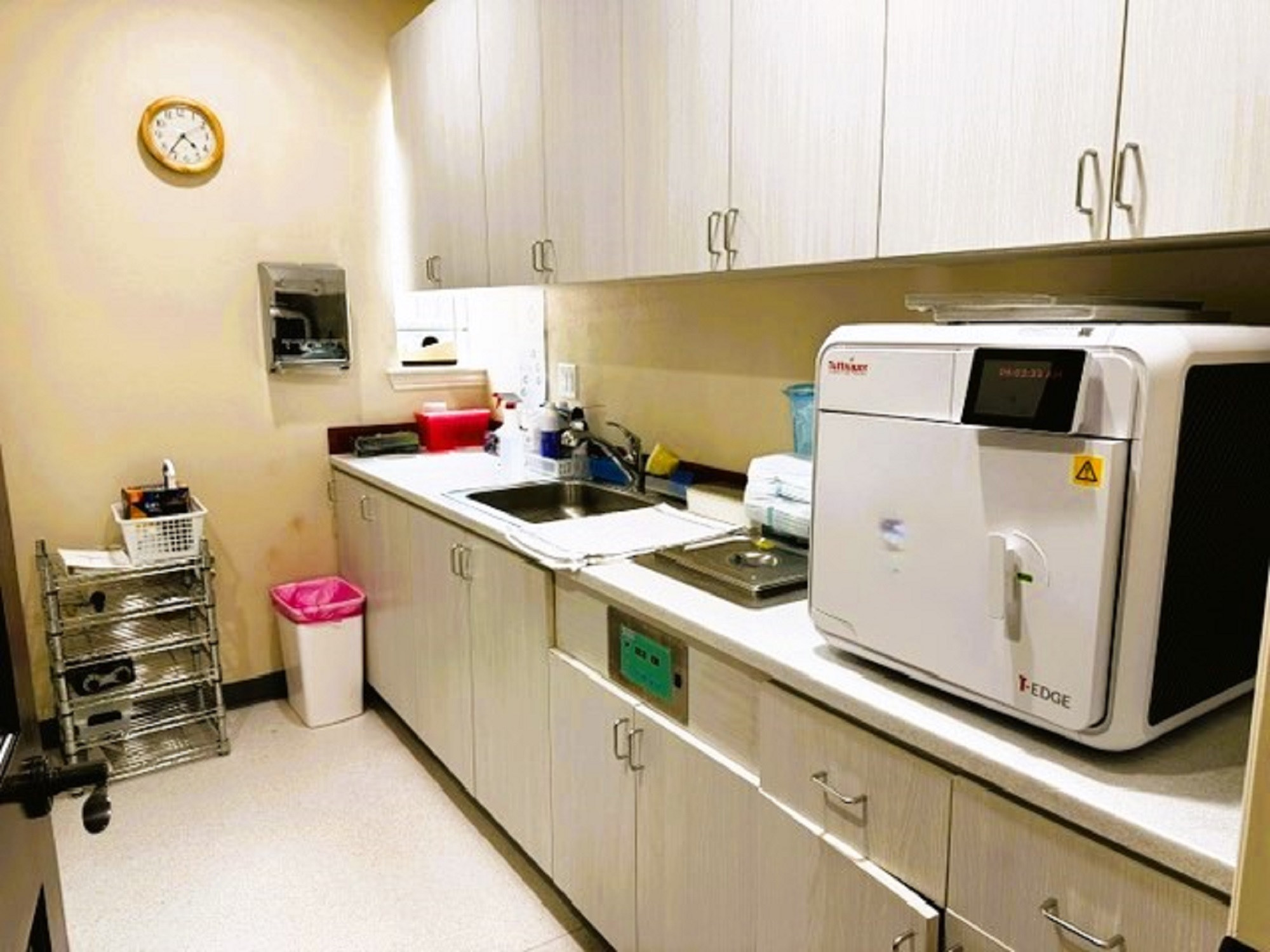 Beige walls with black baseboards and light grey tile flooring. Matching grey upper and lower cabinets line one entire wall with a matching countertop that holds lots of dental sterilization equipment.