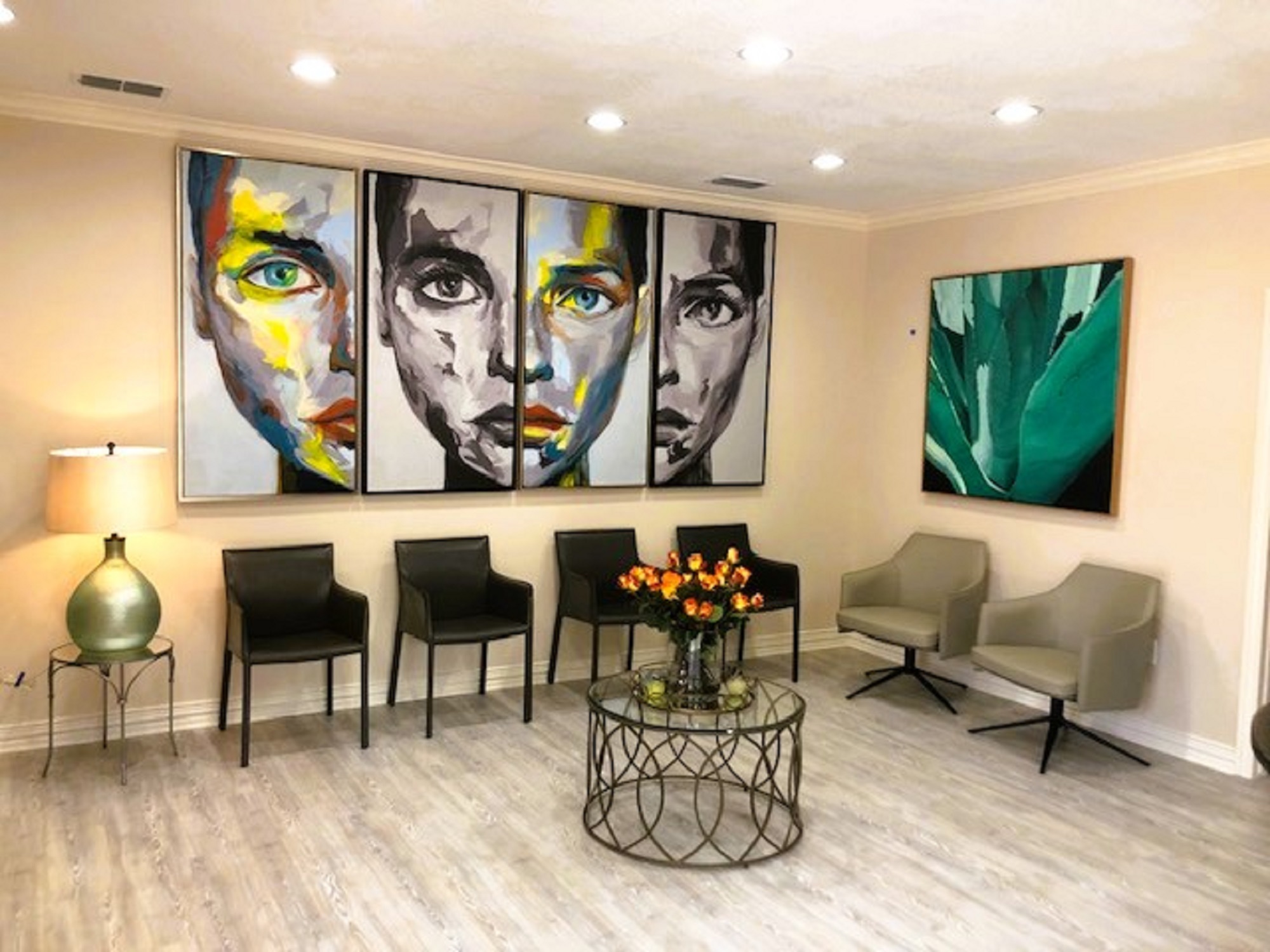 Beige walls with cream crown molding and tall base boards. Grey stained wood look flooring. Multiple can lights light up the over sized artwork on the walls. Several chairs sit along the wall under the art with an end table holding a large lamp. There's a round coffee table sitting in the middle of the room.