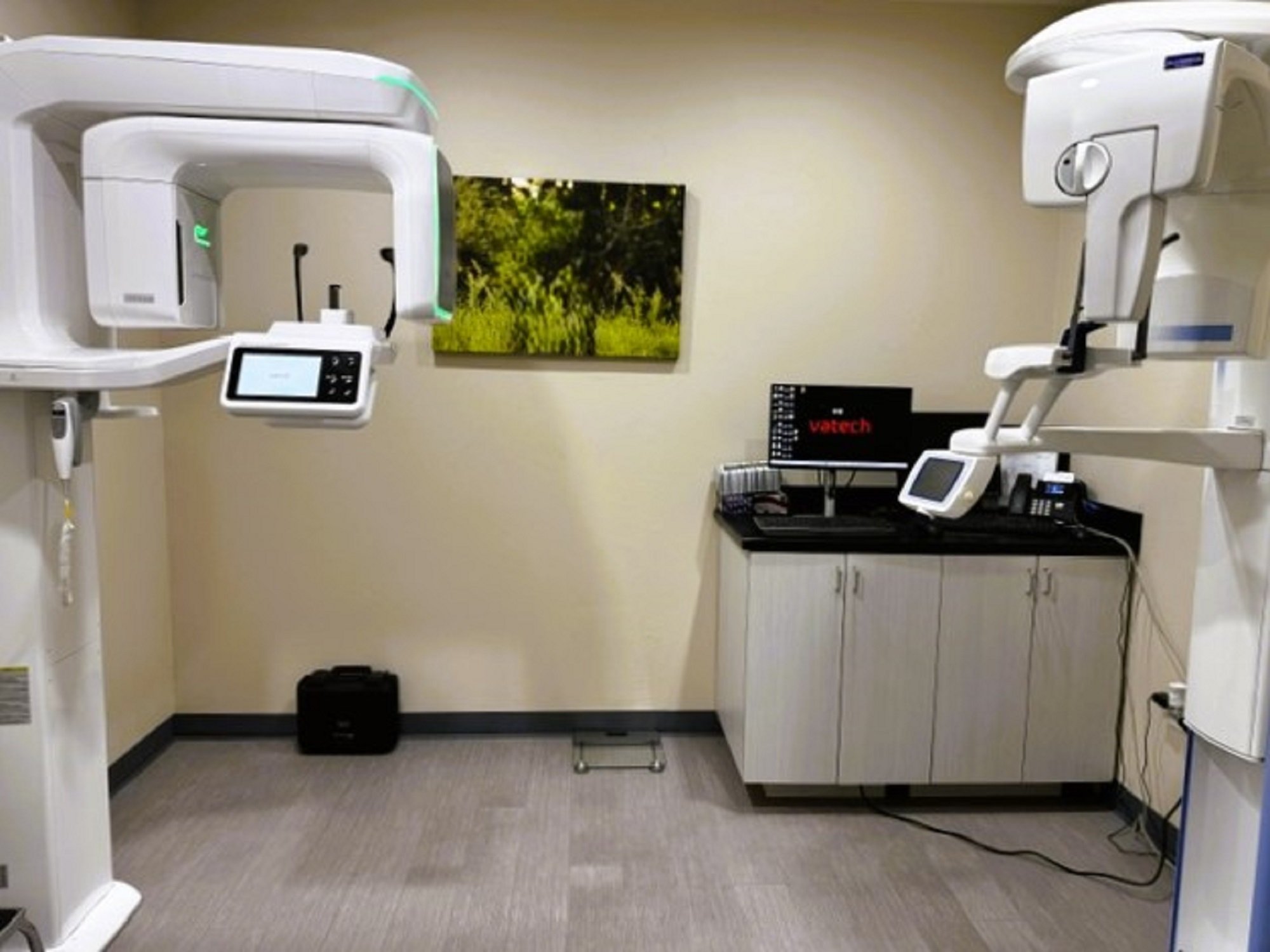 Beige walls with black baseboards and grey tile flooring. Two dental x-ray machines sit opposite each other in the room against either wall.