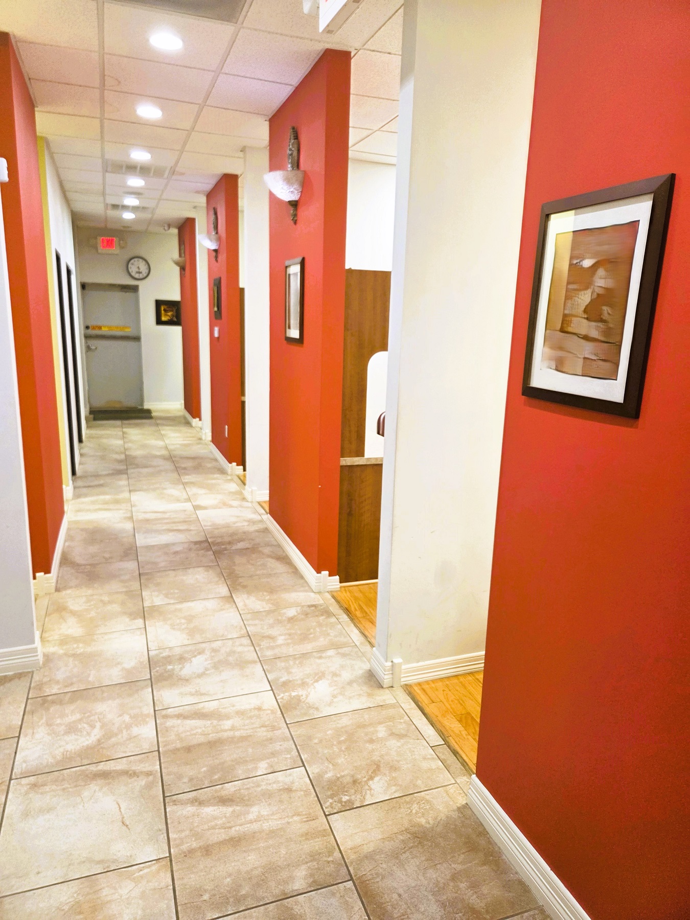 Drop ceiling tiles with can lighting. Alternating red and tan walls with cream trim. Large oversized swirl brown and cream tile flooring go down a long hallway that leads to the different ops, offices, lab, and x-ray.