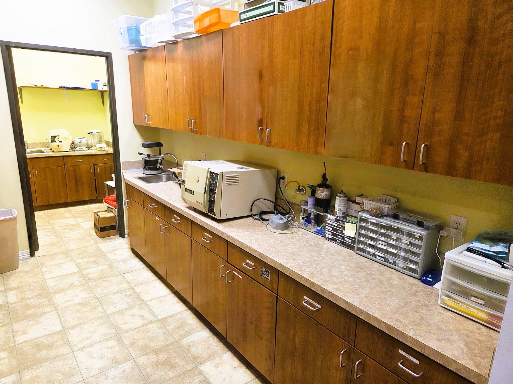 Light grey walls with black door jam and large brown and cream swirled tile flooring. Built in frame-less cabinetry hangs the full length of the wall both uppers and lowers with light colored countertops holding lots of dental lab tools, equipment, and supplies. and a stainless steel single basin sink.