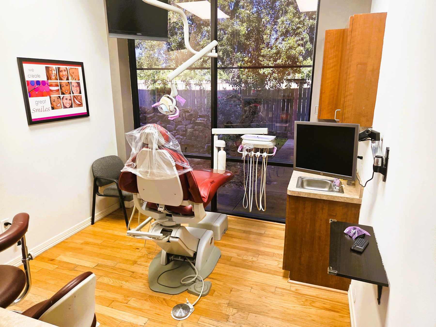 light grey walls with white baseboard. Floor to ceiling window looking out into a quiet yard with a tree. A brown dental procedural chair anchors the room with matching rolling chairs for the dentist and an assistant and an overhead dental light. Natural wood plank flooring and stained wood cabinetry with light countertops hold all the different tools and equipment needed.
