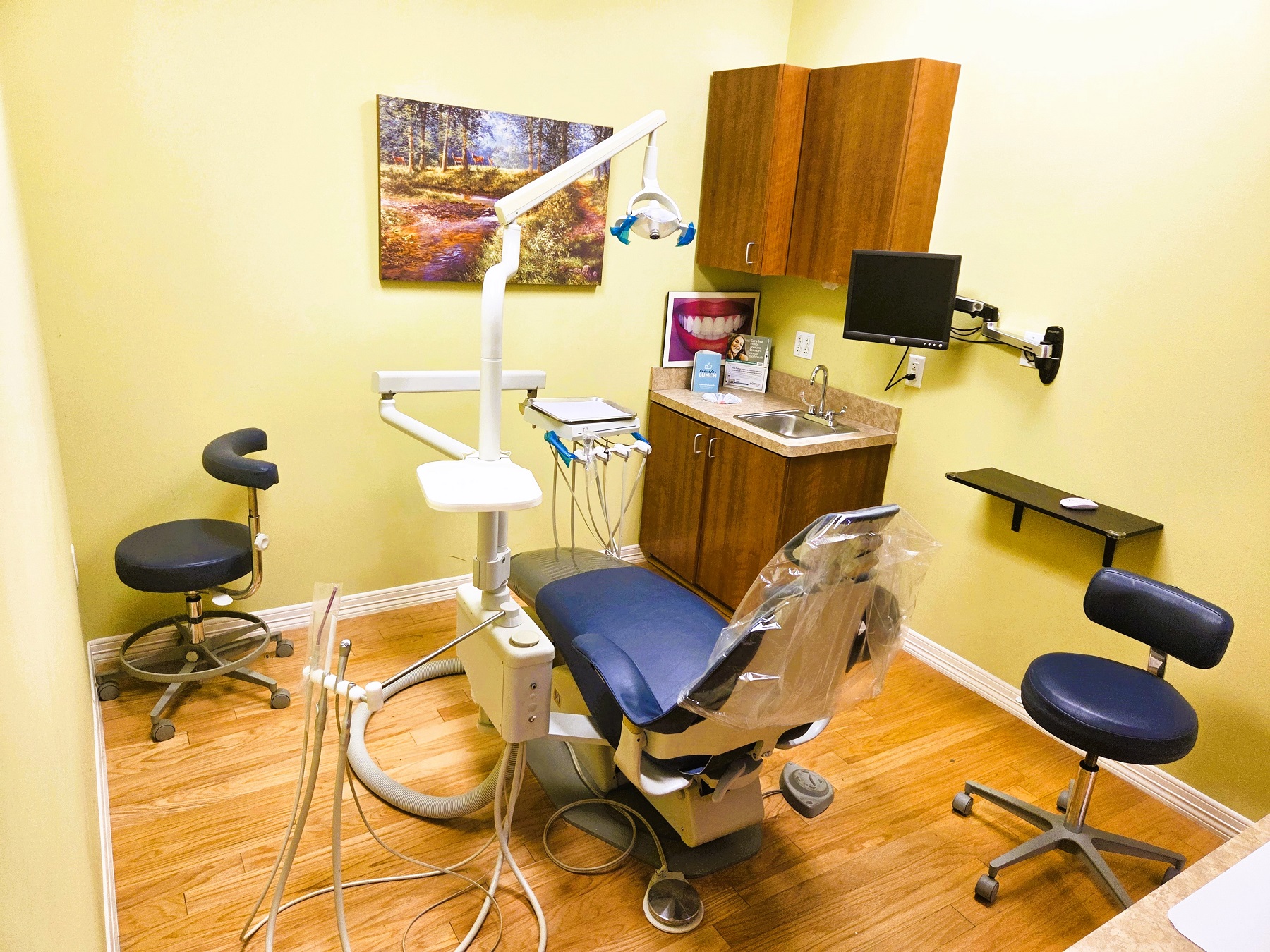 light colored walls with white baseboard. A dark blue dental procedural chair anchors the room with matching rolling chairs for the dentist and an assistant and an overhead dental light. Natural wood plank flooring and stained wood cabinetry with light countertops hold all the different tools and equipment needed. A piece of artwork hangs on the wall