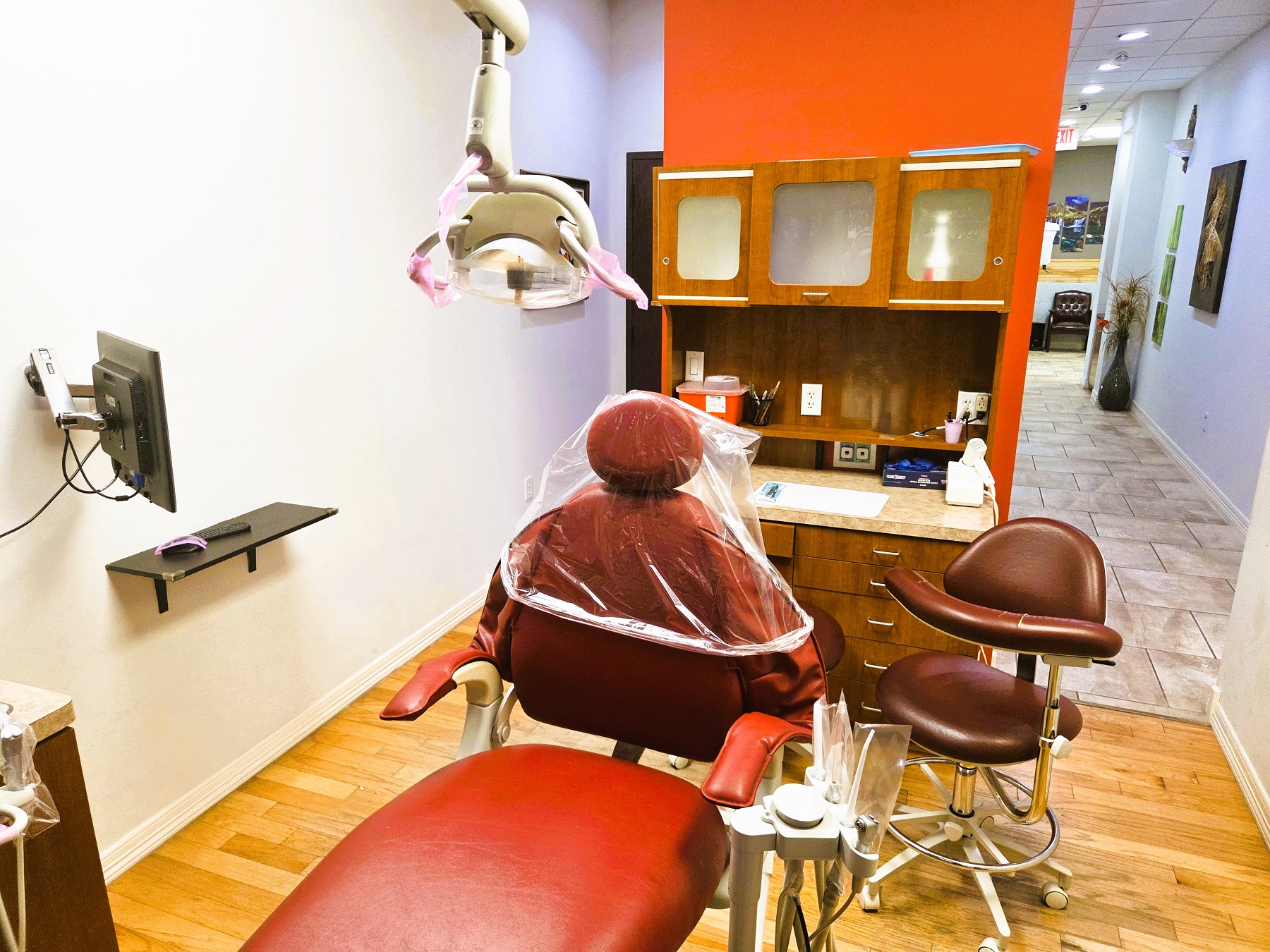 light grey walls with white baseboard. A dark burnt orange dental procedural chair anchors the room with matching rolling chairs for the dentist and an assistant and an overhead dental light. Natural wood plank flooring and burnt orange cabinetry with light countertops hold all the different tools and equipment needed. A flat screen monitor and shelf hang on the wall to the left.