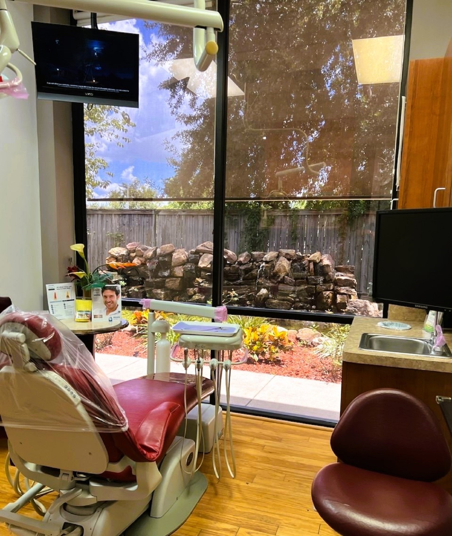 light grey walls with white baseboard. Floor to ceiling window looking out into a quiet courtyard with a pond. A brown dental procedural chair anchors the room with matching rolling chairs for the dentist and an assistant and an overhead dental light. Natural wood plank flooring and stained wood cabinetry with light countertops hold all the different tools and equipment needed.