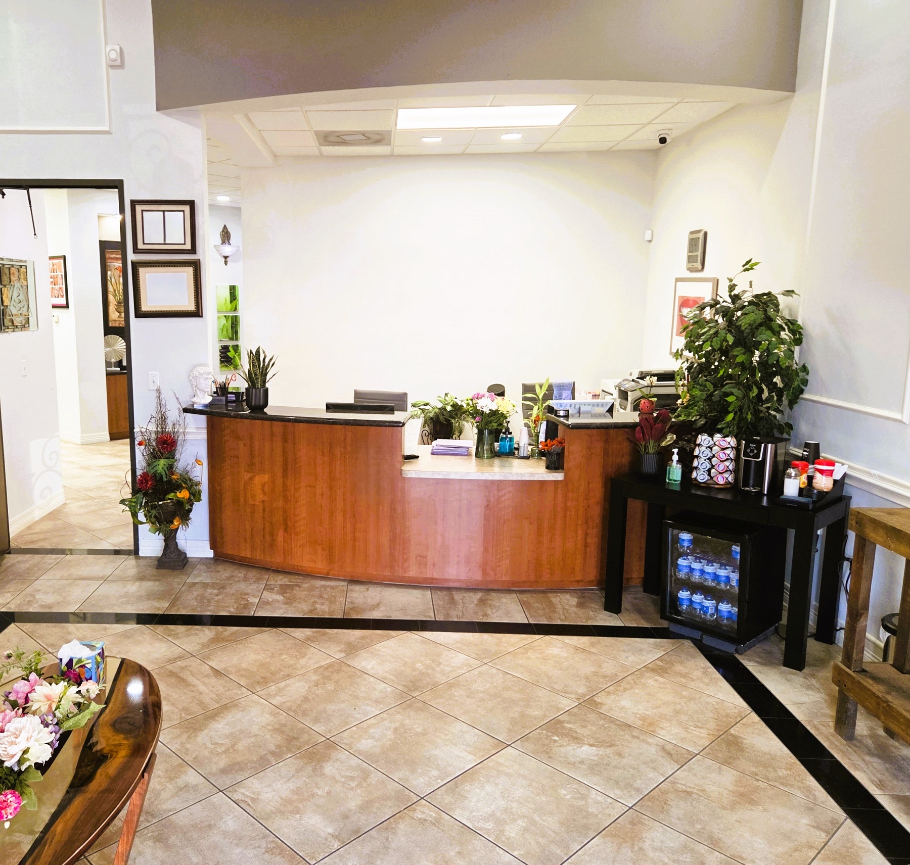 White walls with black trim work and parquet flooring. A medium stained curved desk with a black stone countertop sits in the corner. Lots of plants, artwork, and plaques are around the reception desk. A coffee bar/welcome station sits immediately to the right of the desk to provide refreshments and both hot and cold drinks while they wait in the spacious waiting area.