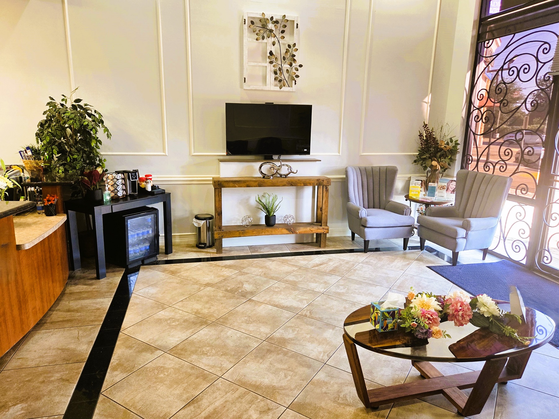Light colored wall with cream chair-rail, baseboard, and wainscotting. Natural wood parquet flooring with a black framing detail in the floor. The entry door has a blue floor mat sitting inside it and several cushioned red leather chairs with wooden arms surround a coffee tale and there's an end table with a plant in the corner. The coffee table has lots of plants and flowers on it.