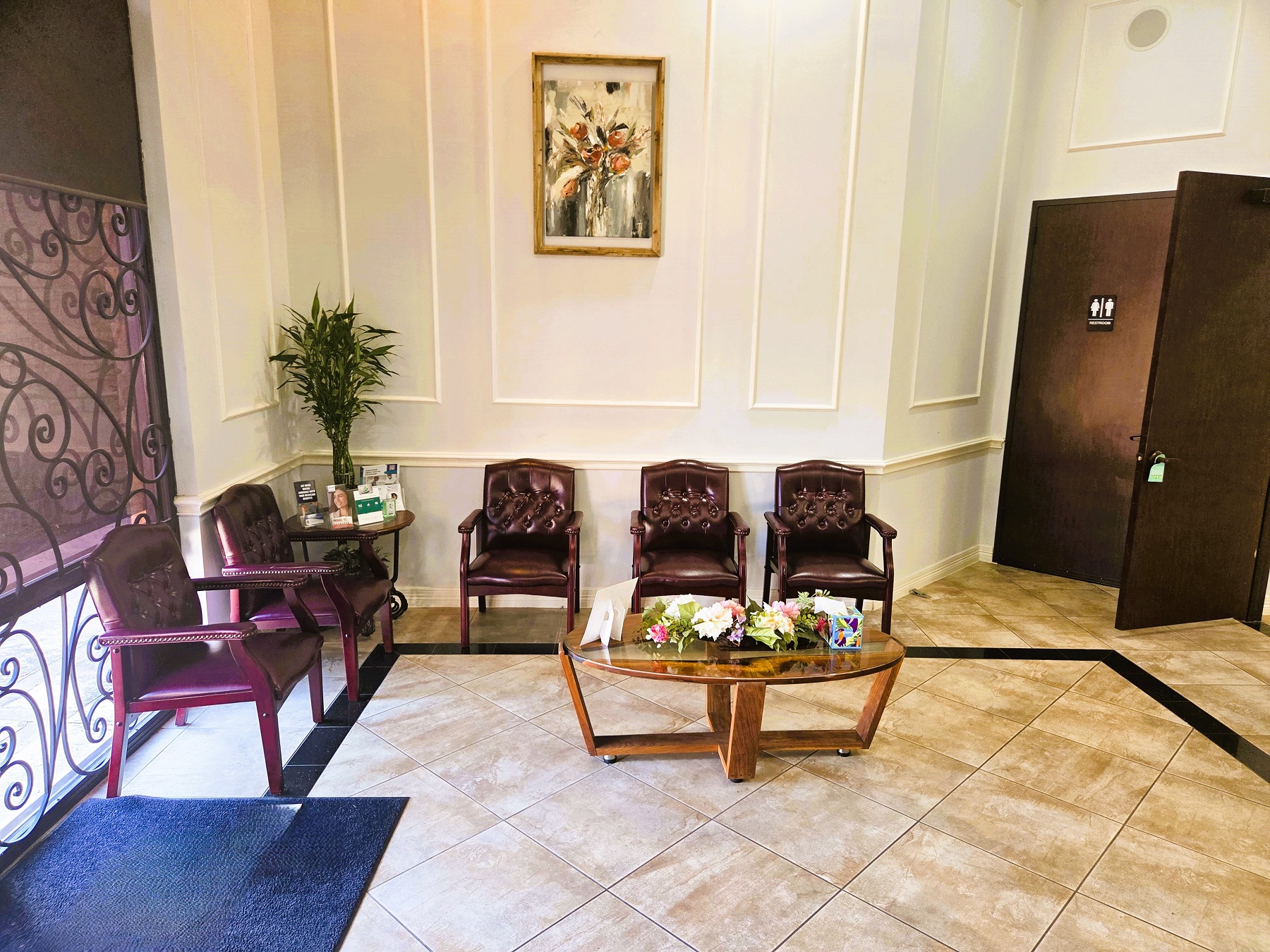 Light colored wall with cream chair-rail, baseboard, and wainscotting. Natural wood parquet flooring with a black framing detail in the floor. The reception desk is to the left with it's refreshment center. A flat-screen tv sits on a side table against the wall and a pair of overstuffed arm chairs sit around an end table in the corner.