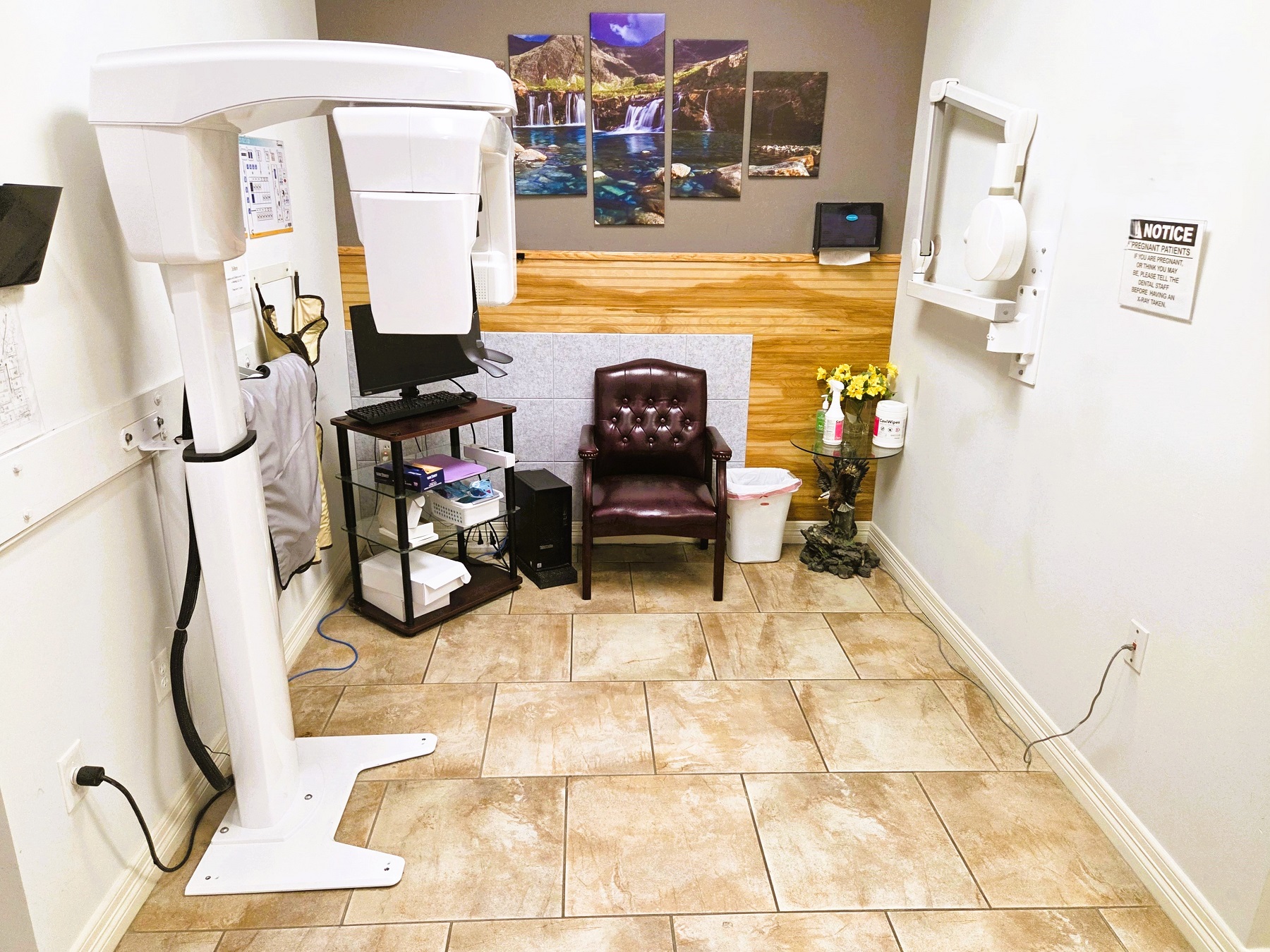 Light grey walls with white baseboard and large offset brown and cream swirl tile flooring. Accent wall in the back with photo artwork and wood details. A cushioned leather chair sits against the back wall and a rolling cart with a monitor sits in the corner. A large dental x-ray machine sits against the wall.