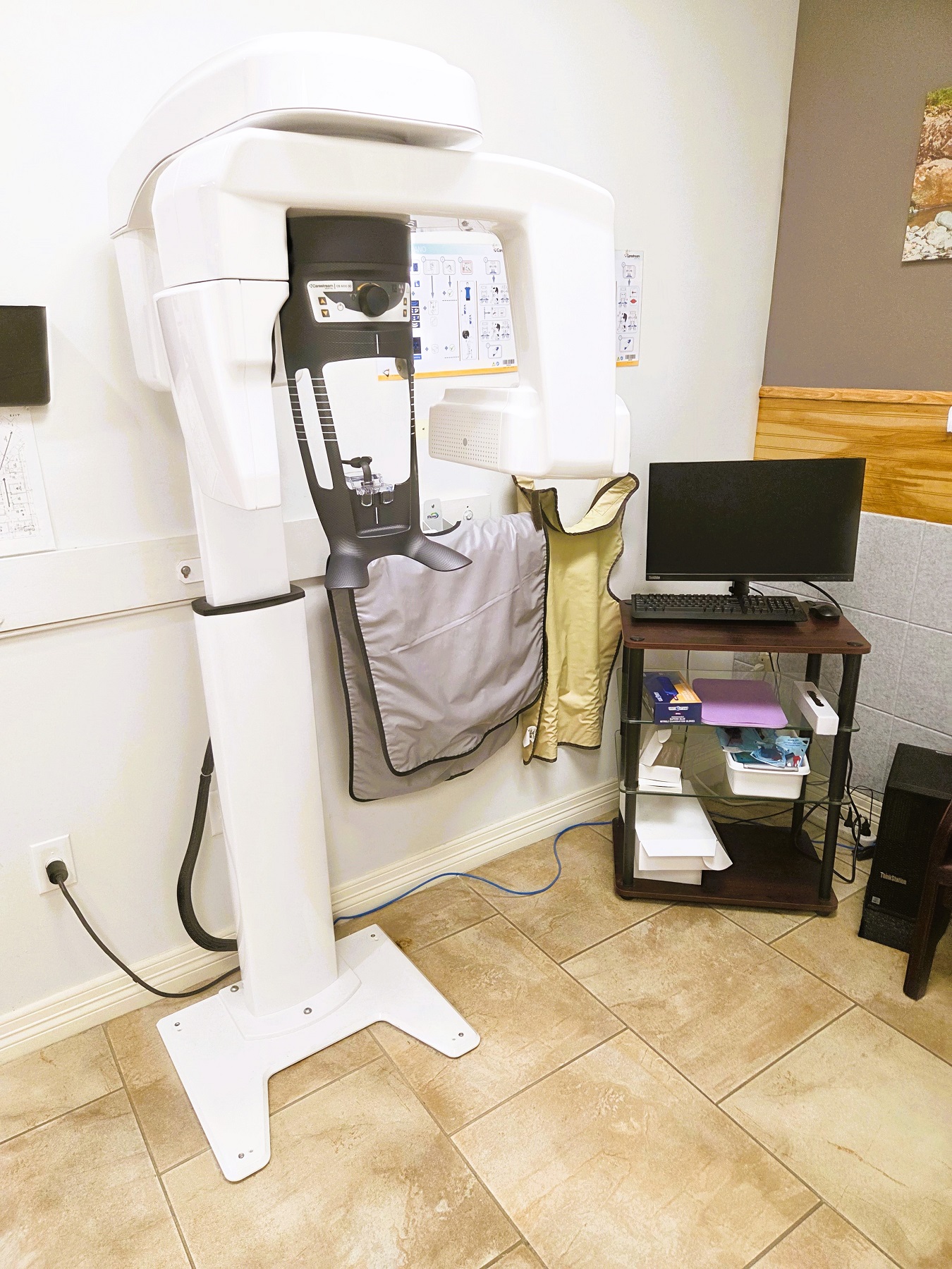Light grey walls with white baseboard and large offset brown and cream swirl tile flooring. Accent wall in the back with photo artwork and wood details. A cushioned leather chair sits against the back wall and a rolling cart with a monitor sits in the corner. A large dental x-ray machine sits against the wall.