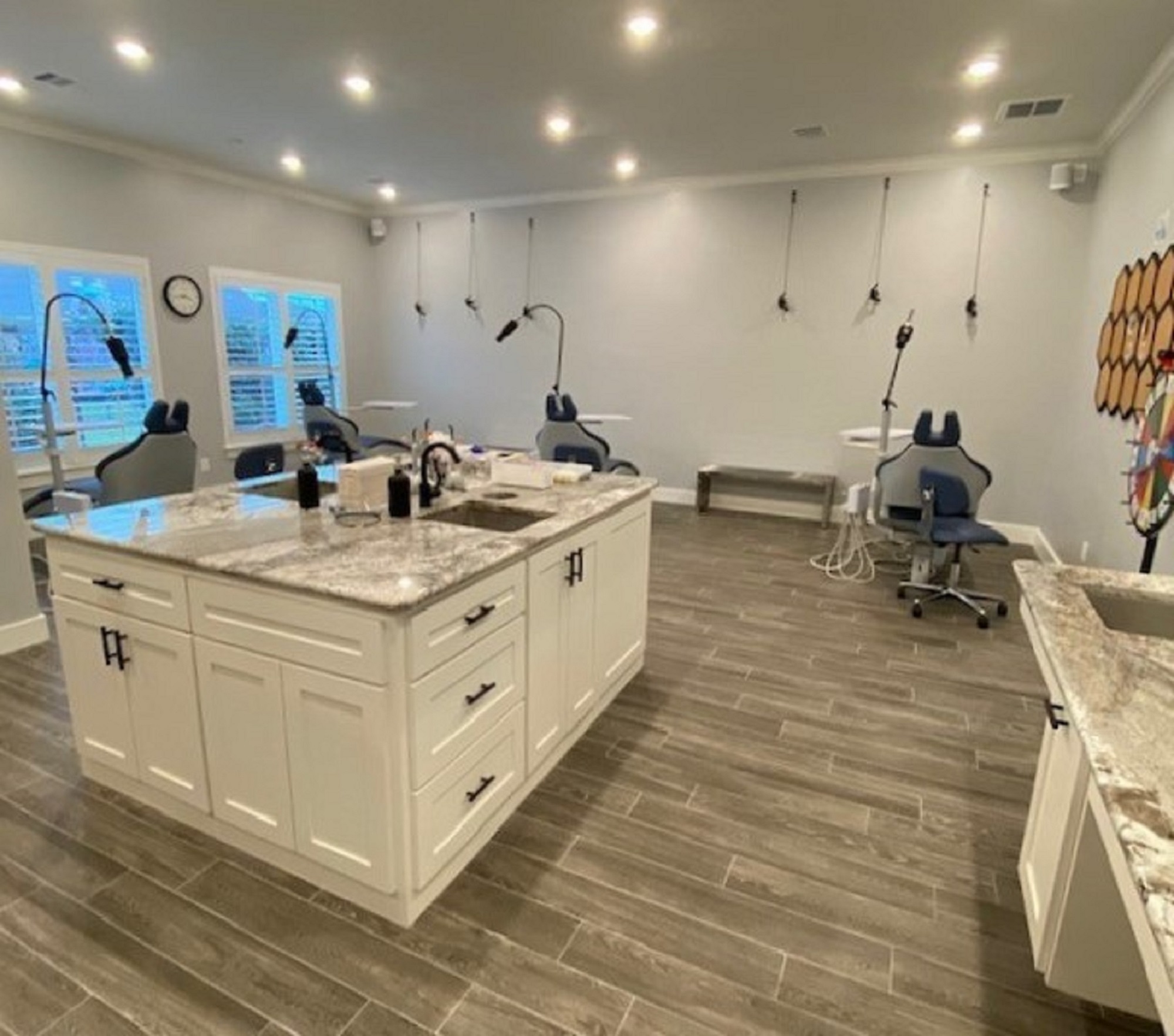 Light grey walls with white crown molding, casement windows, and baseboards. Grey wood-look flooring planks. A huge white island with a white and grey marble countertop and two sinks holds lots of equipment. A matching cabinet and sink are on the right-hand wall as well. 4 dental procedural chairs surround two sides of the island. Lots of can lights and dental procedural lights provide tons of lighting in the room.