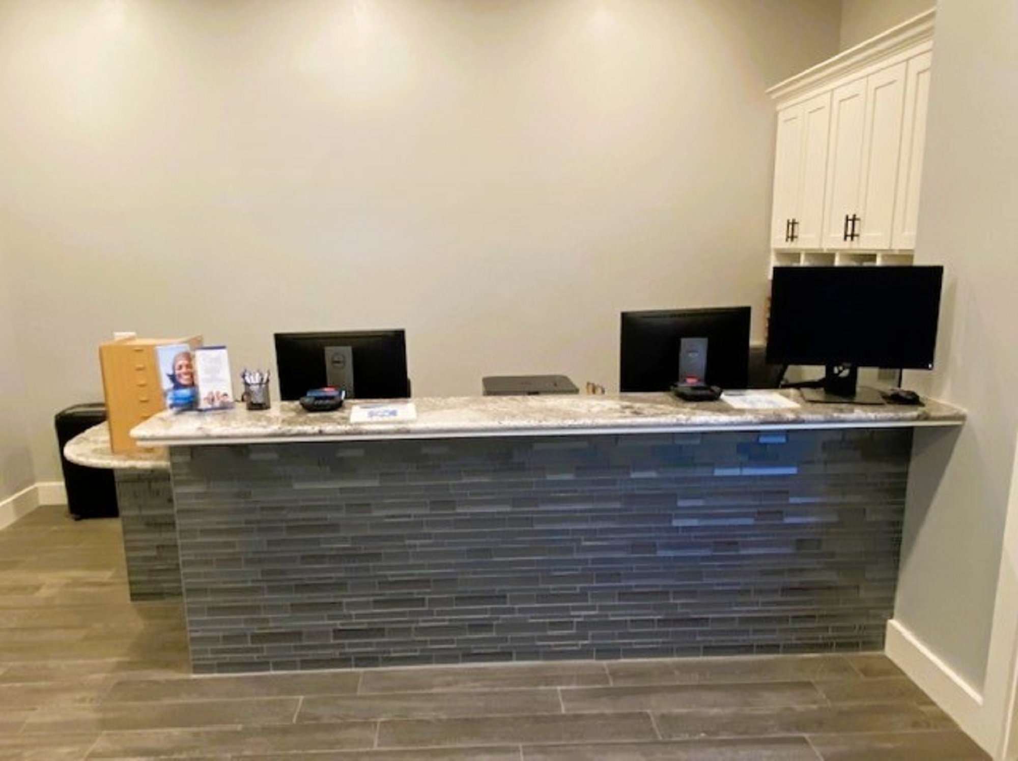 Light grey walls with white baseboards. Grey wood-look flooring and a large builtin desk with a stacked grey stone front and white and grey marble stone desk top that holds lots of office equipment and supplies. Behind the desk and on the right is a white builtin cabinet to hold office supplies.