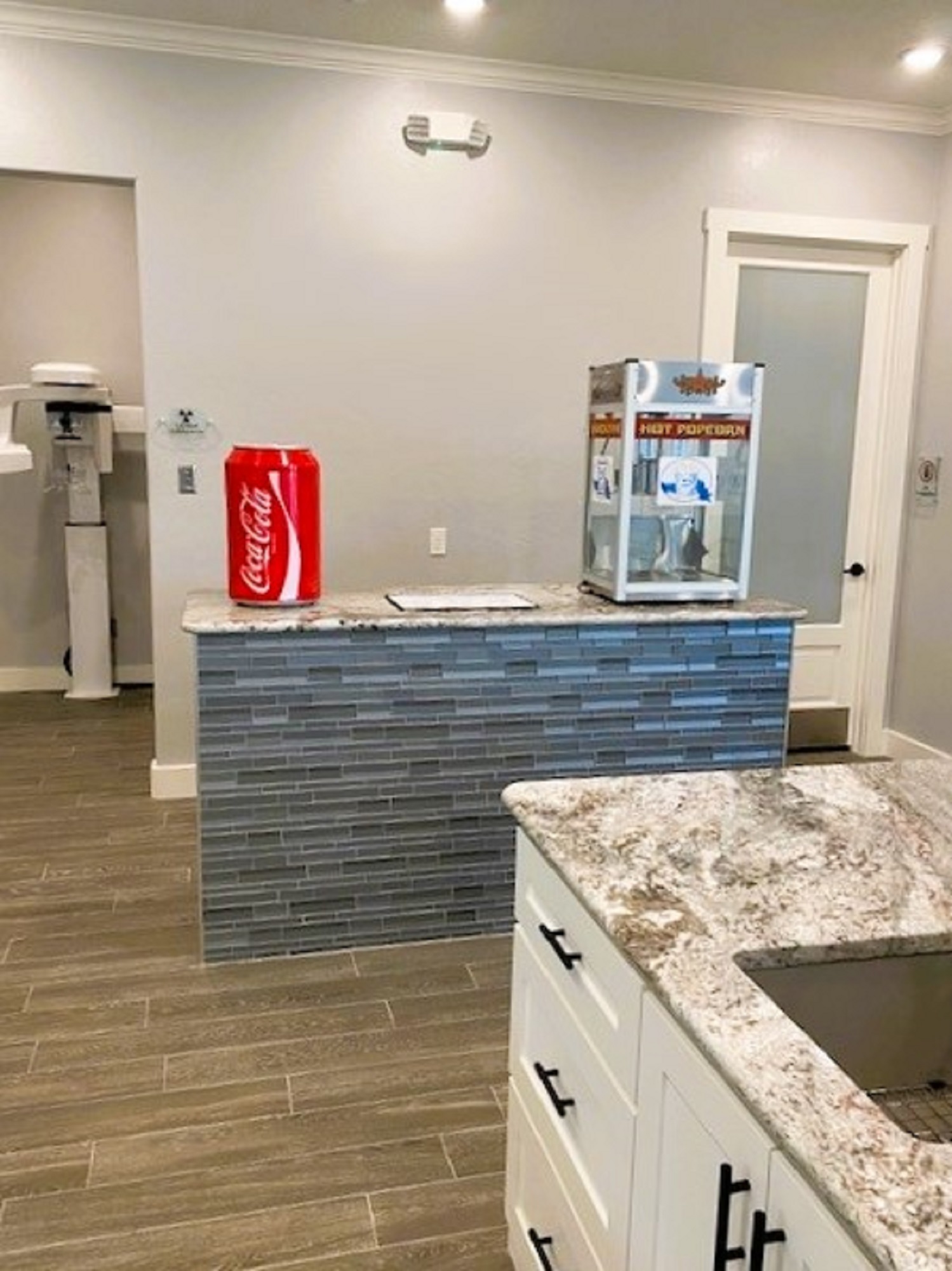 Light grey walls with white crown molding, door trim, and baseboards. Grey wood-look plank flooring. A medium size island with tile sides and a white and grey marble stone top has a giant Coca Cola can on the counter top along with a popcorn machine. A dental z-ray machine can be seen through the door way to the left of the refreshment station.
