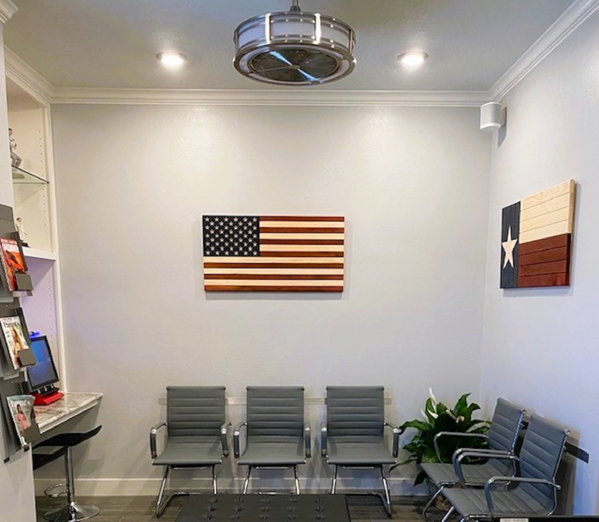 Light grey walls with white crown molding and baseboard and grey commercial carpet on the floor. Grey cushioned upholstery chairs with stainless steel arms and legs line the room against the walls. On the left-hand wall there is a built-in white bookcase with built-in reading desk and a small stool. There's a wooden American flag on the back wall and a similar sized wooden Texas flag on the right-hand wall. Can lights provide lots of lighting along with a ceiling fan/light.