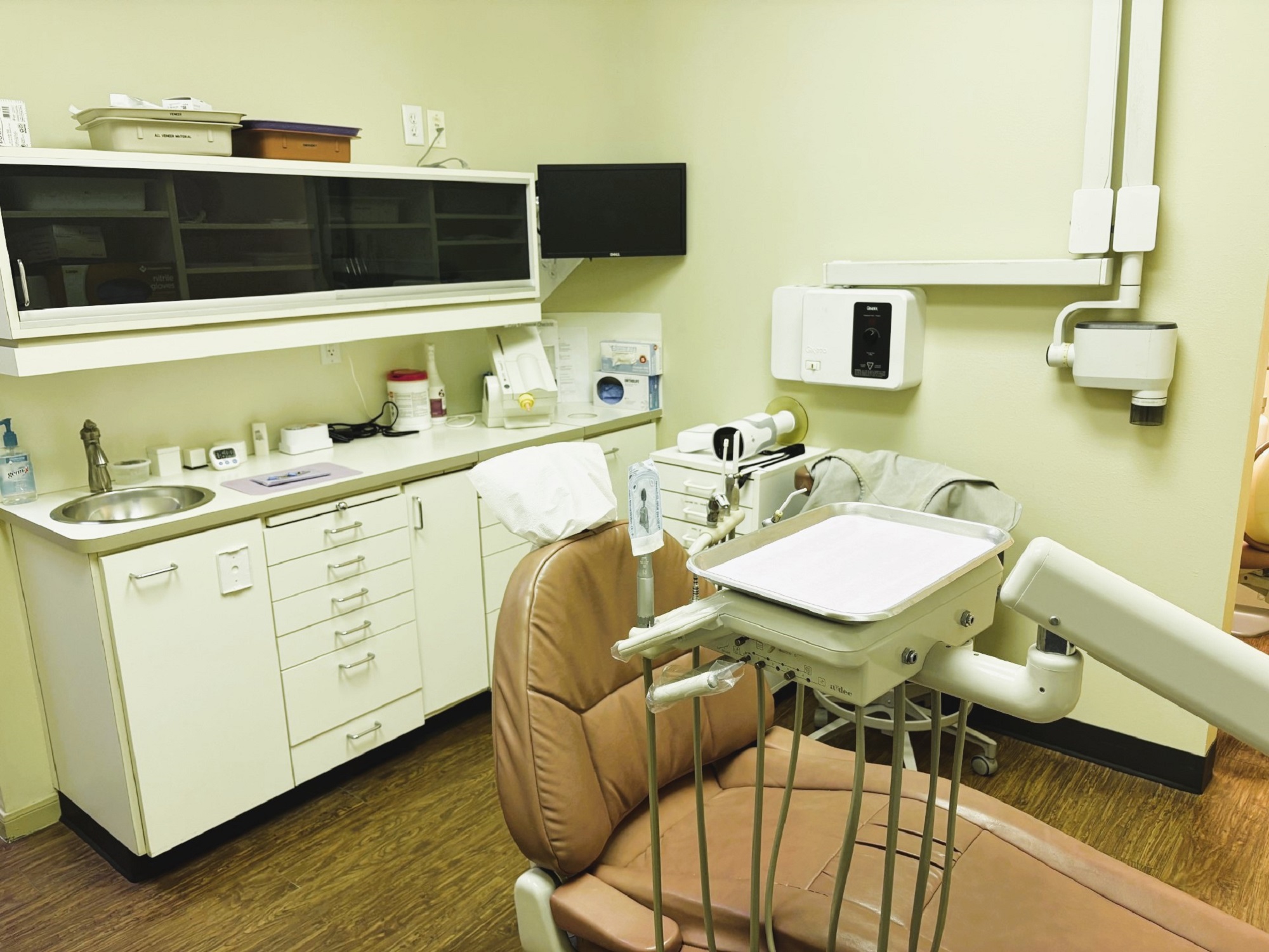 Light colored walls with matching baseboards. Medium stained hardwood flooring. A large, tan cushioned dental procedural chair sits in the middle of the room with overhead dental lighting. There is white cabinetry on the walls with a stainless steel sink in the lower cabinet and lots of space for equipment, tools, and supplies. An x-ray machine is mounted on the adjacent wall.