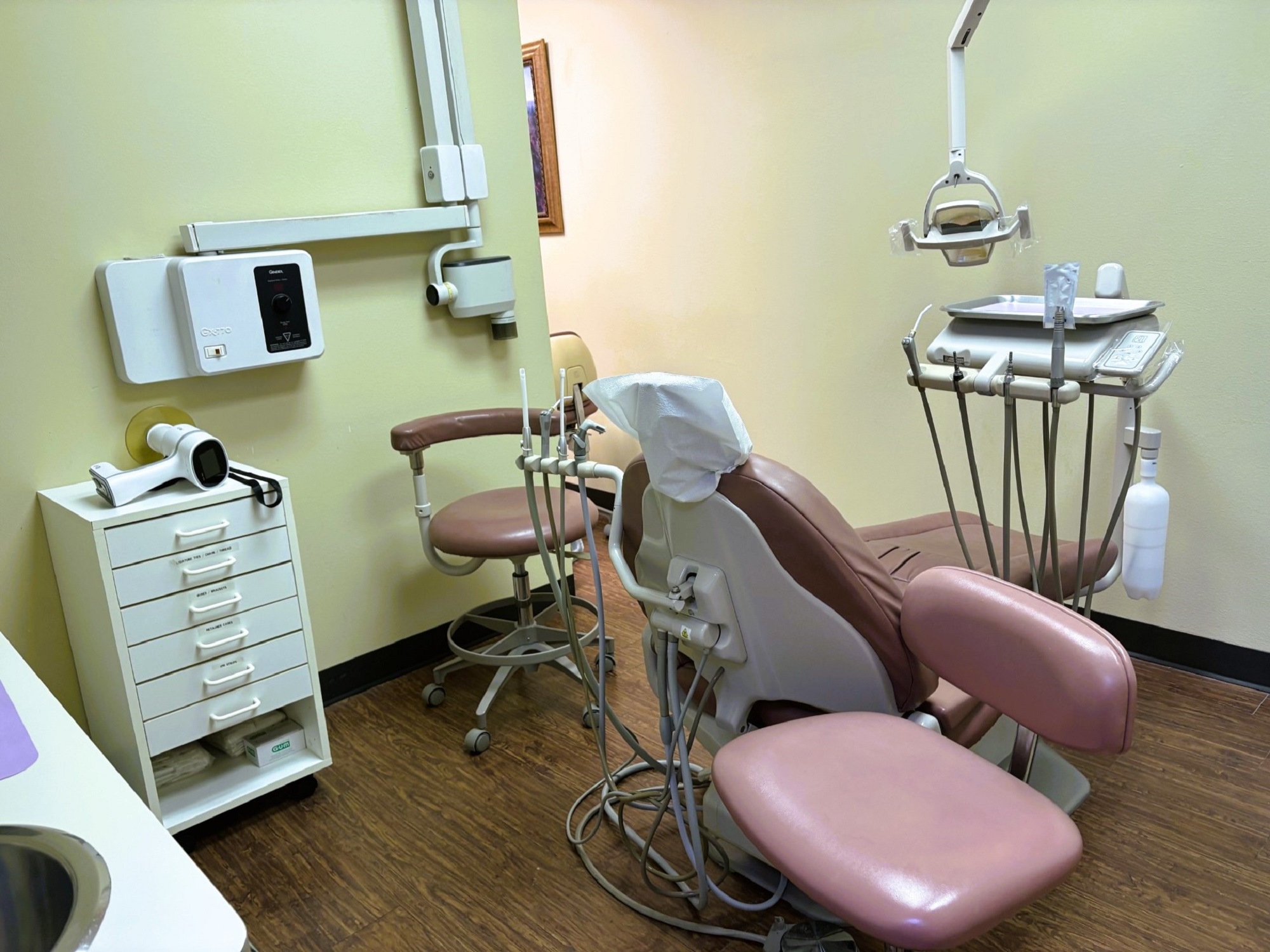 Light colored walls with matching baseboards. Medium stained hardwood flooring. A large, tan cushioned dental procedural chair sits in the middle of the room with overhead dental lighting. There is white cabinetry on the walls with a stainless steel sink in the lower cabinet and lots of space for equipment, tools, and supplies. An x-ray machine is mounted on the adjacent wall.