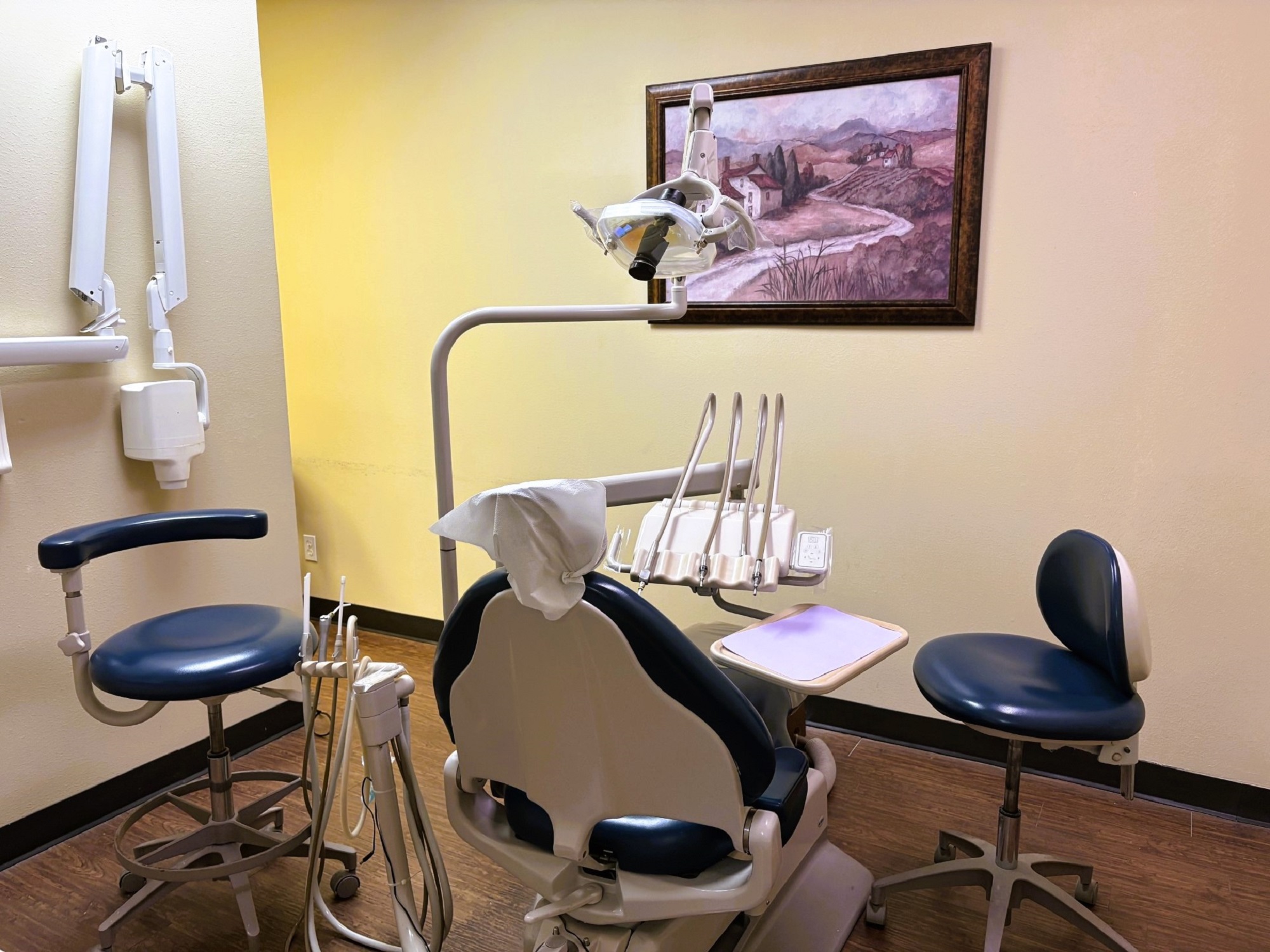 Light colored walls with matching baseboards. Medium stained hardwood flooring. A large, tan cushioned dental procedural chair sits in the middle of the room with overhead dental lighting. There is white cabinetry on the walls with a stainless steel sink in the lower cabinet and lots of space for equipment, tools, and supplies. An x-ray machine is mounted on the adjacent wall.