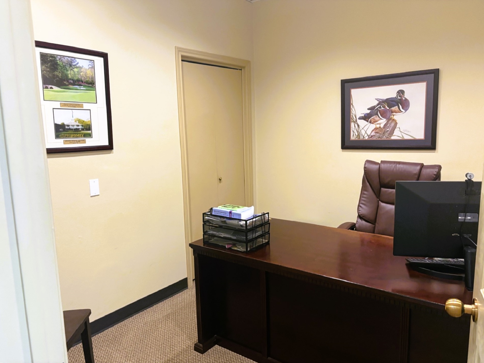 tan walls with dark baseboards and slightly lighter brown commercial carpet flooring. A couple large framed pieces of art hang on the walls A large dark wood desk sits in the middle of the room with a huge leather overstuffed office chair behind it.