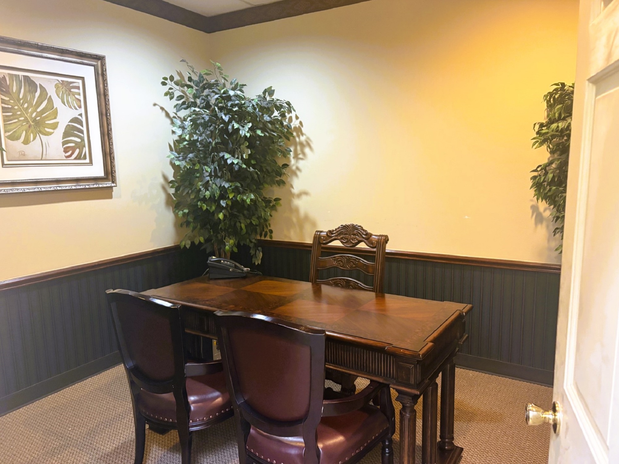Two tone walls with a chair rail. Tan upper walls and dark lower walls with dark baseboards and slightly lighter colored flooring. Large framed artwork hangs on the wall with a couple ficus trees in the corners. A small, dark wood table anchors the room with matching chairs on either side.