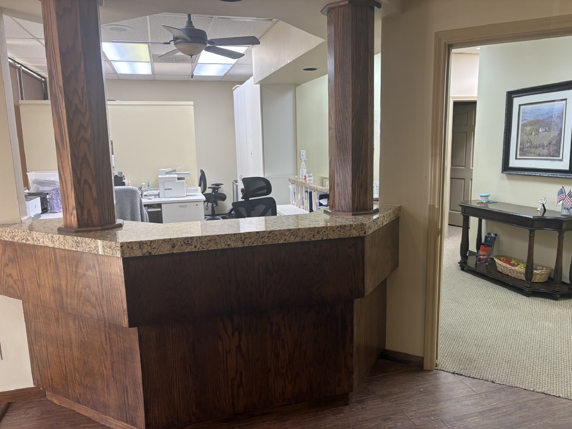 A large, dark wood island built in the middle of the room with dark stone countertops The inside walls and surfaces are painted white with office tools and equipment and dark office chairs. In front of the island are dark hardwood floors leading to tan carpeted flooring in the back.